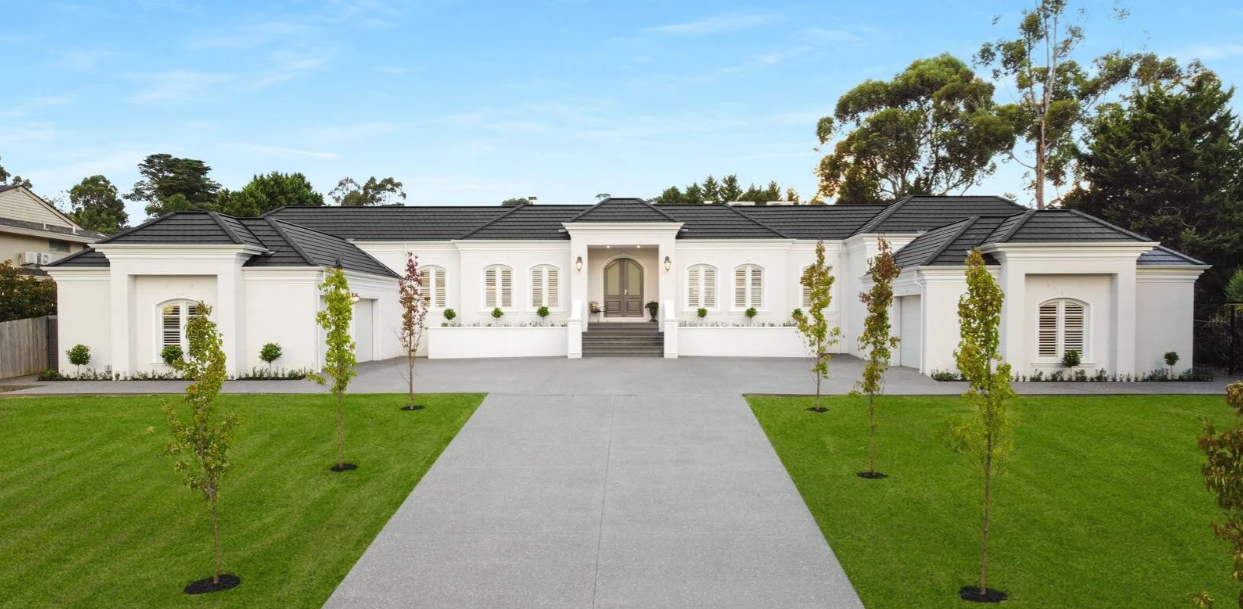 Modern white house with black roof, symmetrical design, front steps, small landscaped front yard with young trees, well-maintained lawn, and blue sky in the background.