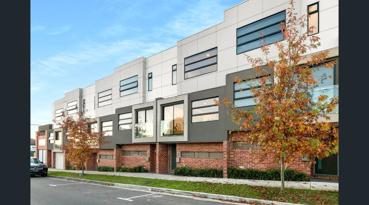 Modern multi-story apartment building with brick and gray exterior, large windows, and trees with autumn foliage in front.