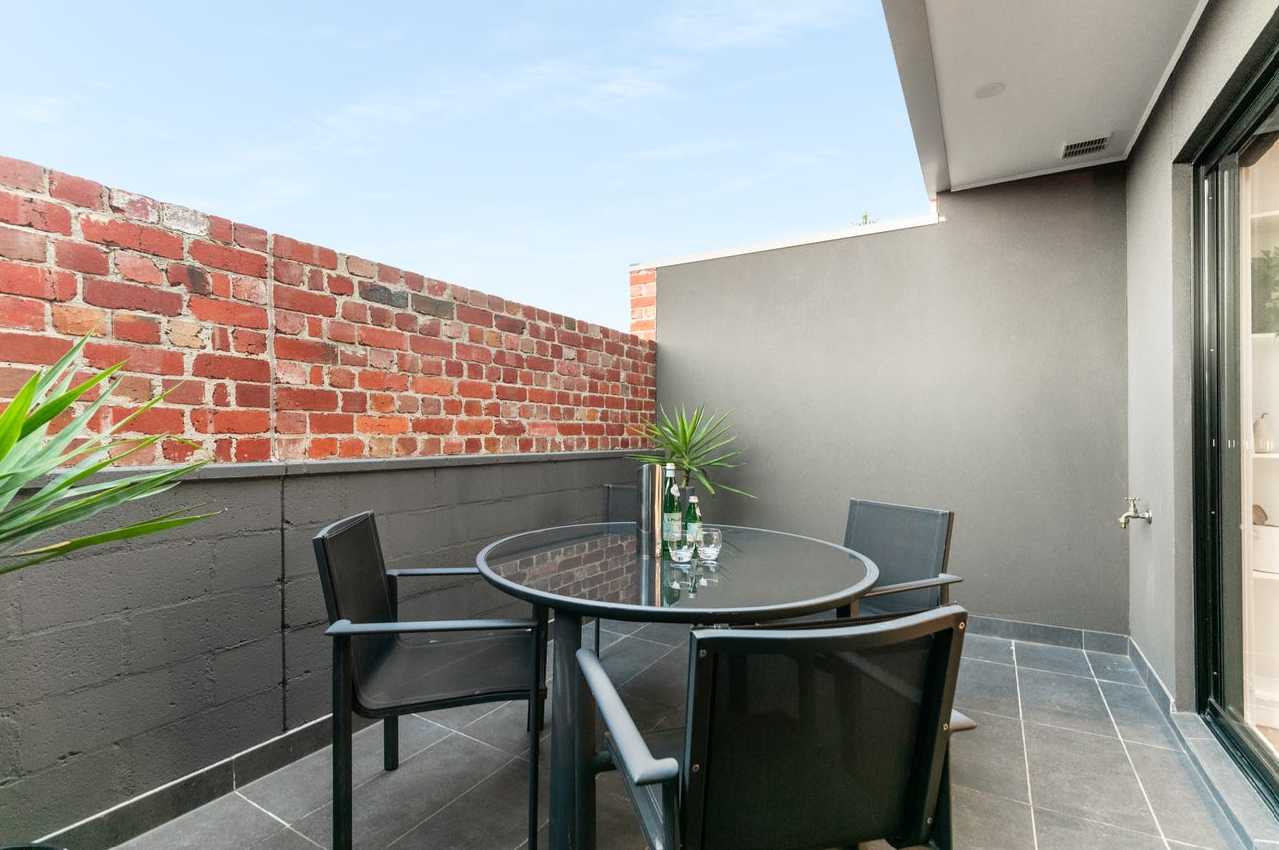 Outdoor balcony with a glass table, four black chairs, a potted plant, and bottled beverages against a brick and concrete wall under a blue sky.