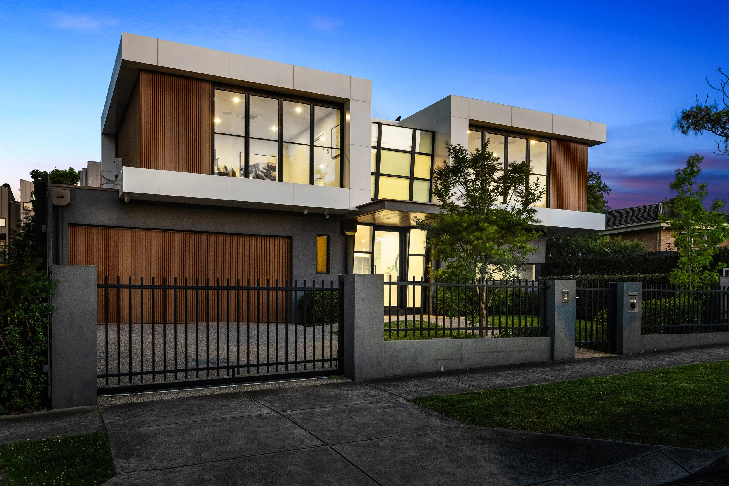 Modern two-story house with large glass windows, wooden accents, a black metal fence, and a green front yard during sunset.