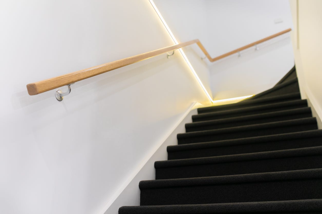 Indoor staircase with black carpeted steps, wooden handrails, and white walls illuminated by soft yellow lighting.