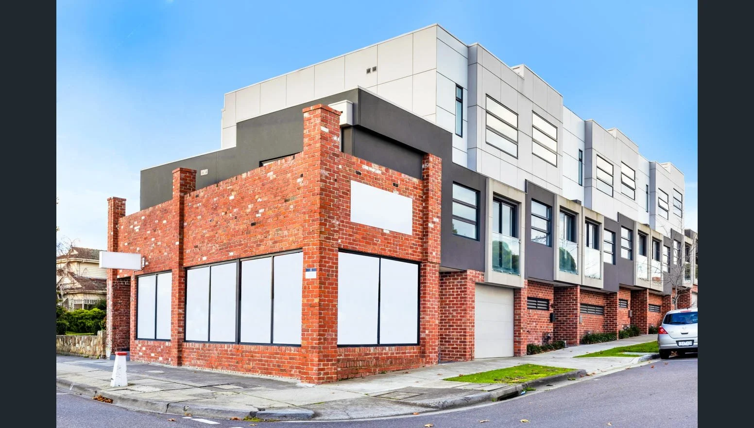 A modern multi-story building with a brick ground floor and upper floors with gray and white siding, surrounded by a sidewalk and a parked car, under a clear blue sky.