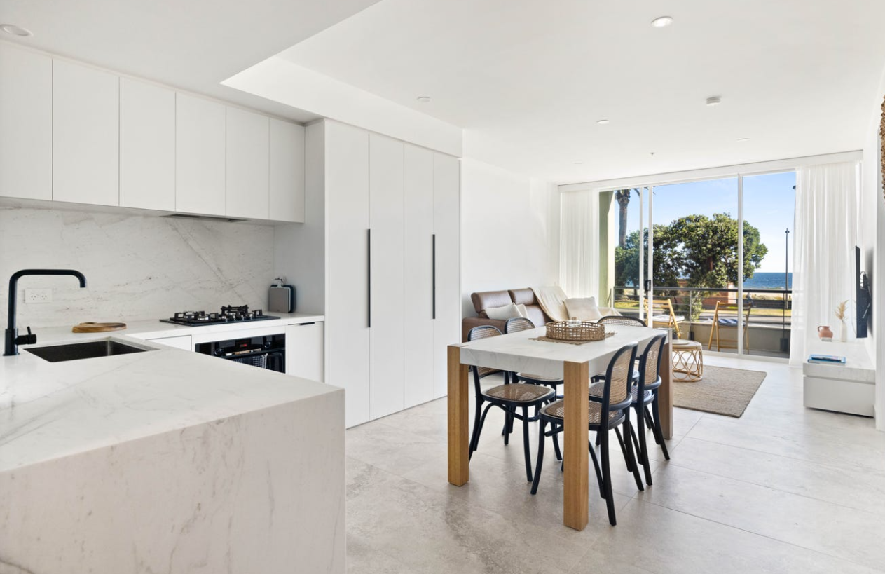 Bright, modern open-concept kitchen and living room with white cabinetry, marble countertops, a dining table, and sliding glass doors leading to a balcony with a view of trees and the ocean.