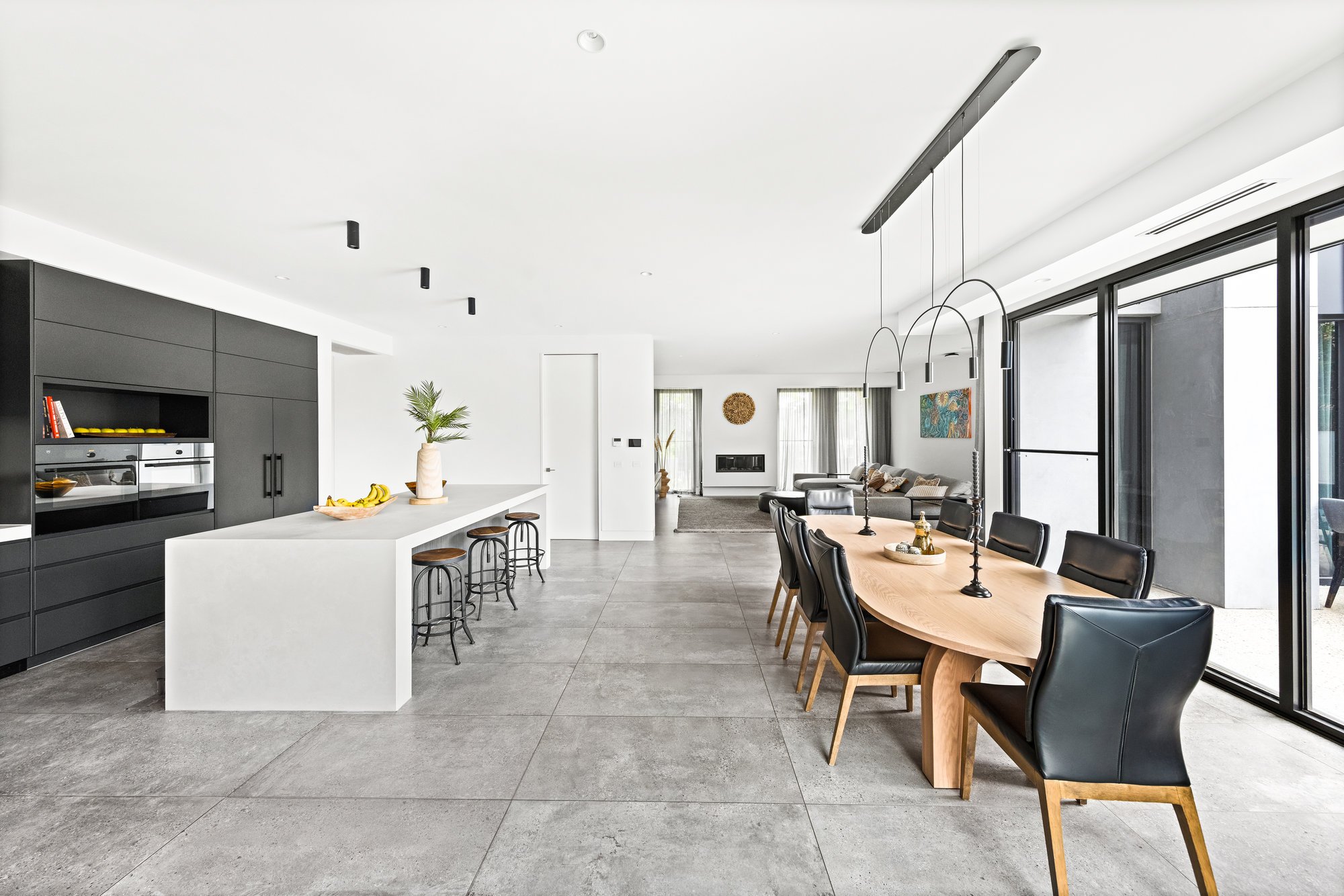 Open-concept living and dining area with black kitchen cabinets, white island, wooden dining table with black chairs, large windows, and a gray sofa in the background.