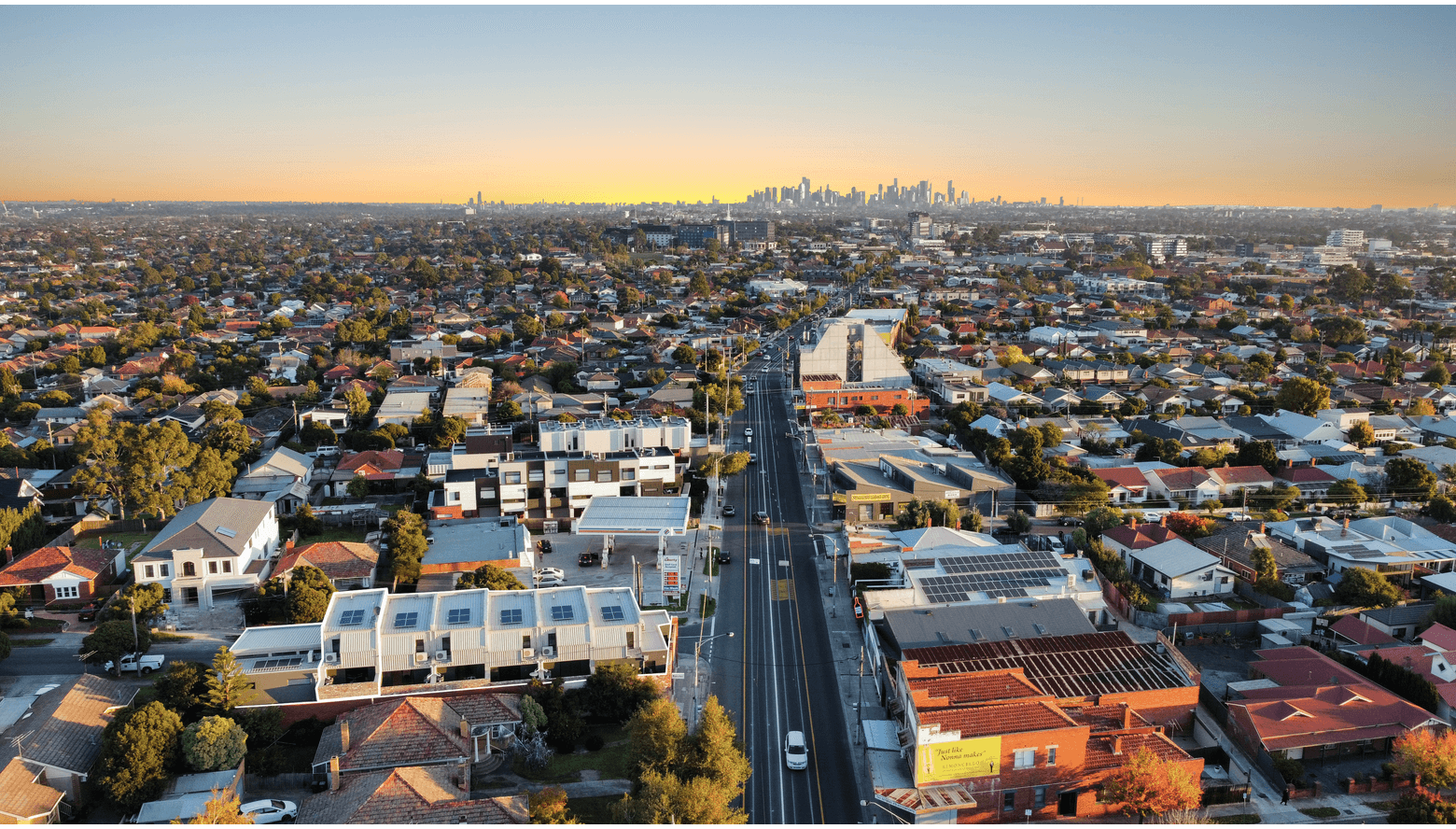 Aerial view of a city neighborhood at sunset with a downtown skyline in the background, featuring residential houses, commercial buildings, and streets.