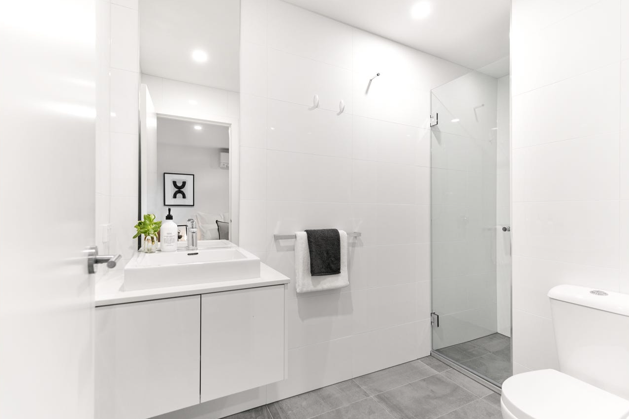 Modern white bathroom featuring a sink with a cabinet, a mirror, a towel rack with a black towel, a glass shower enclosure with a door, a toilet, and minimal decor like a small plant and soap.