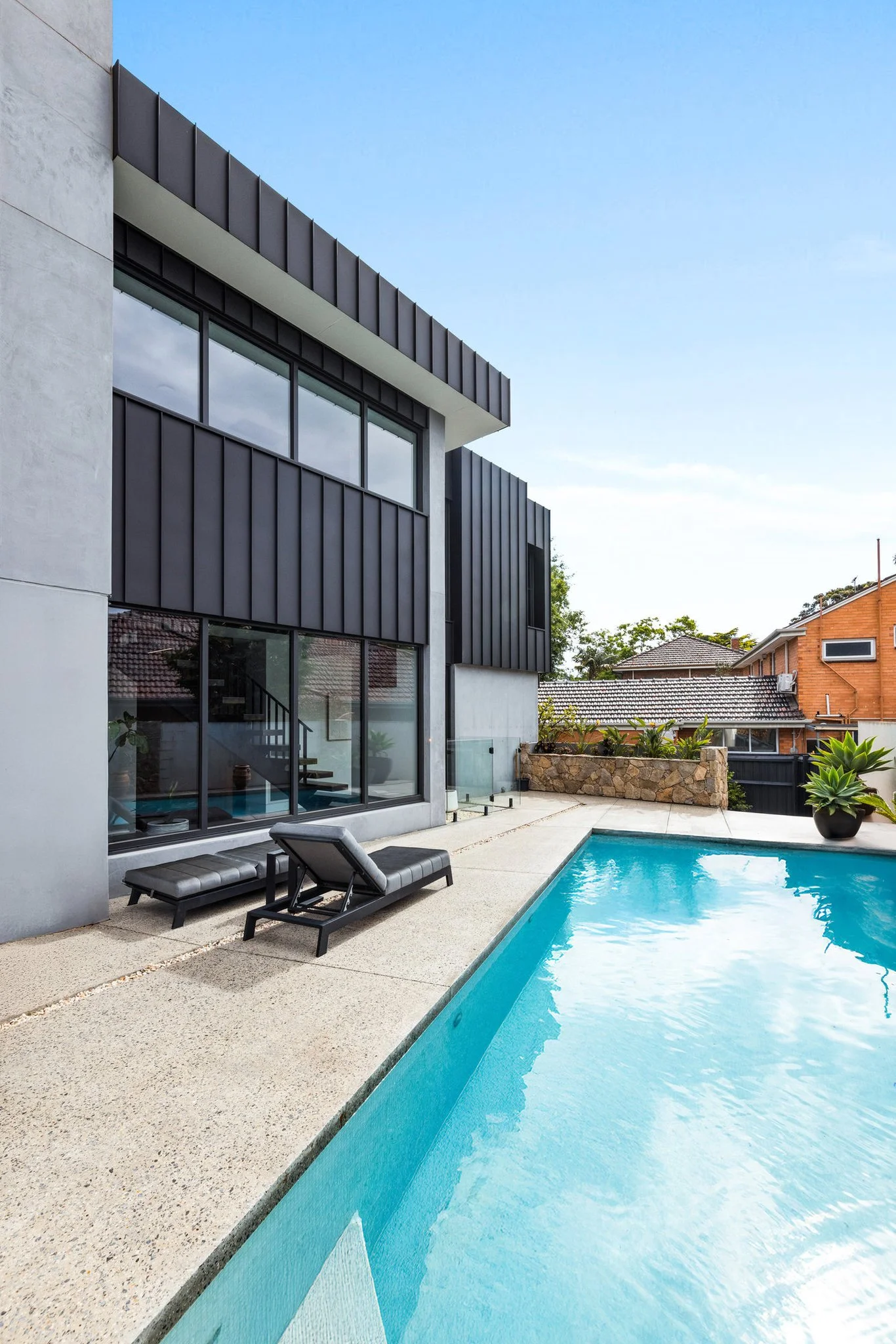 Modern house with large glass windows and black exterior panels, outdoor pool with lounge chairs, and neighboring houses in the background under a blue sky.