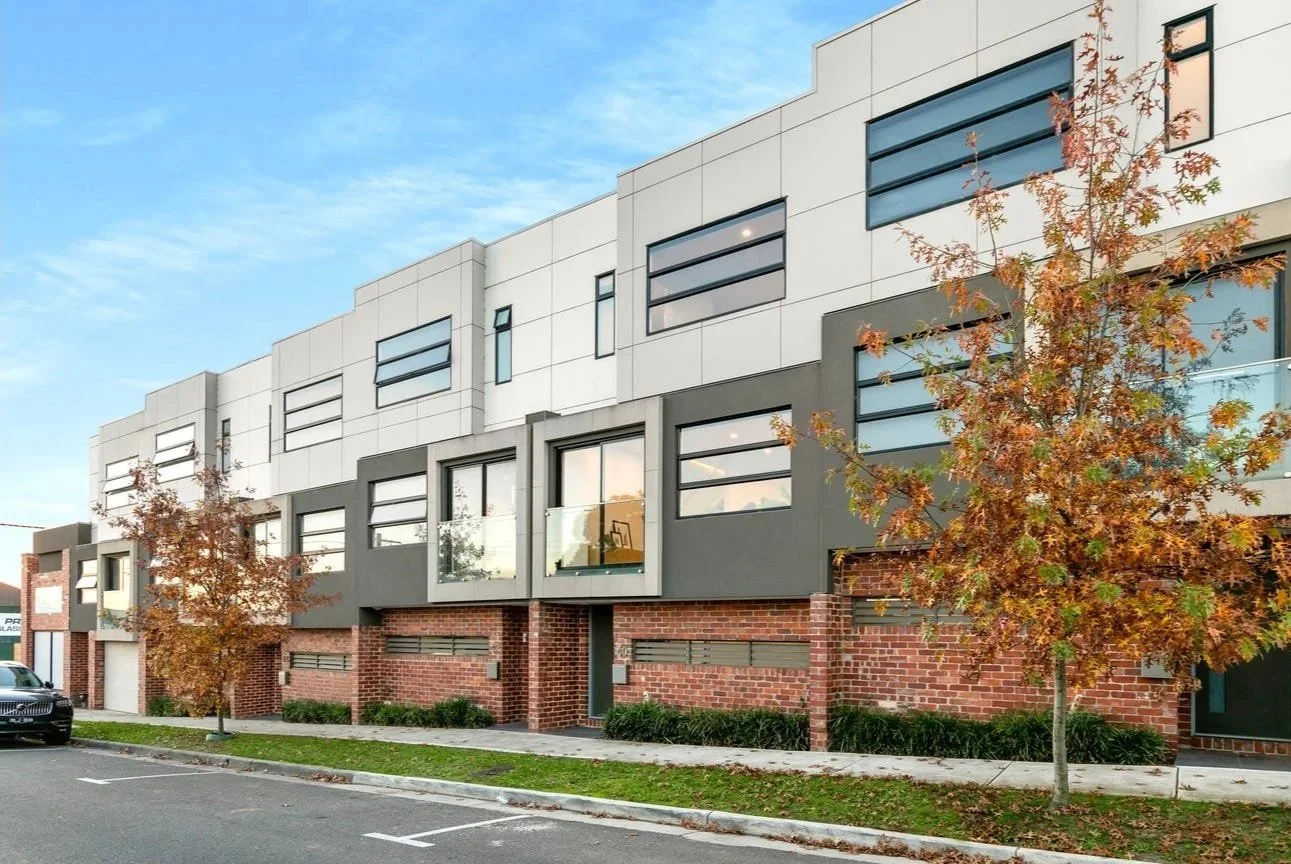 Modern residential building with brick and gray exterior, large windows, and small trees with autumn leaves in front.