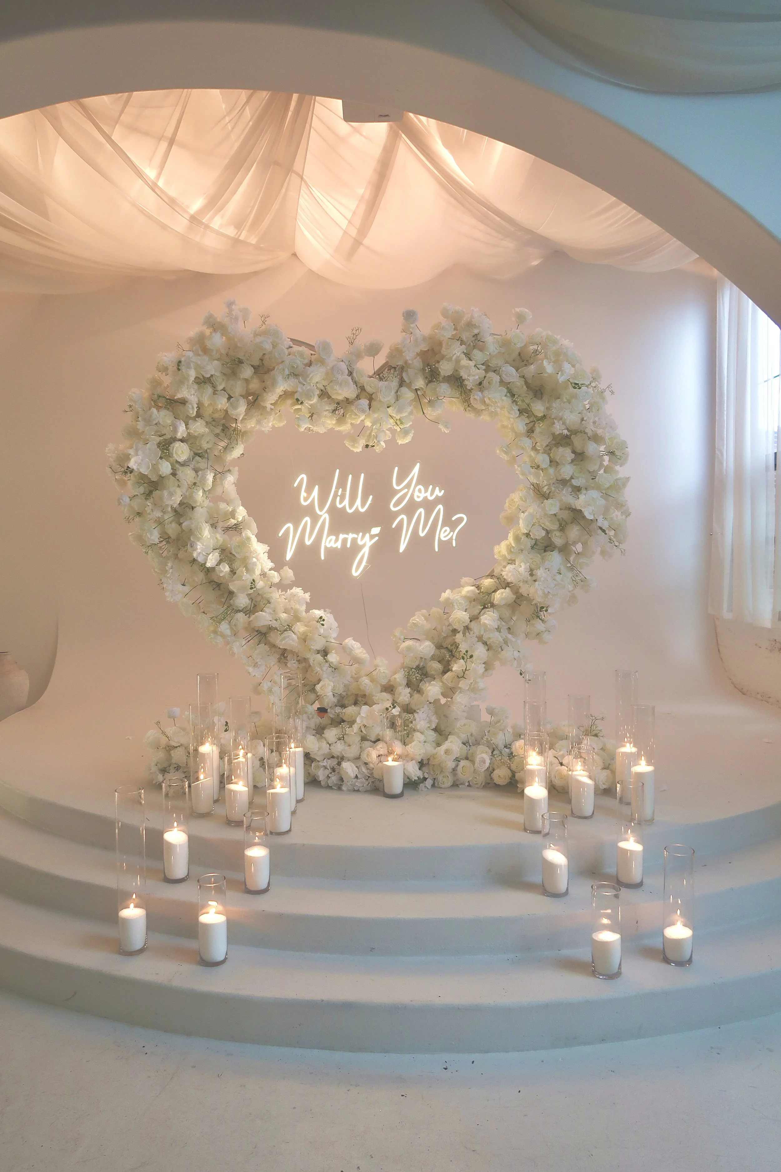Luxury proposal setup with 8 ft white rose heart arch, white sand wax candles and neon 'Marry Me' sign in NYC
