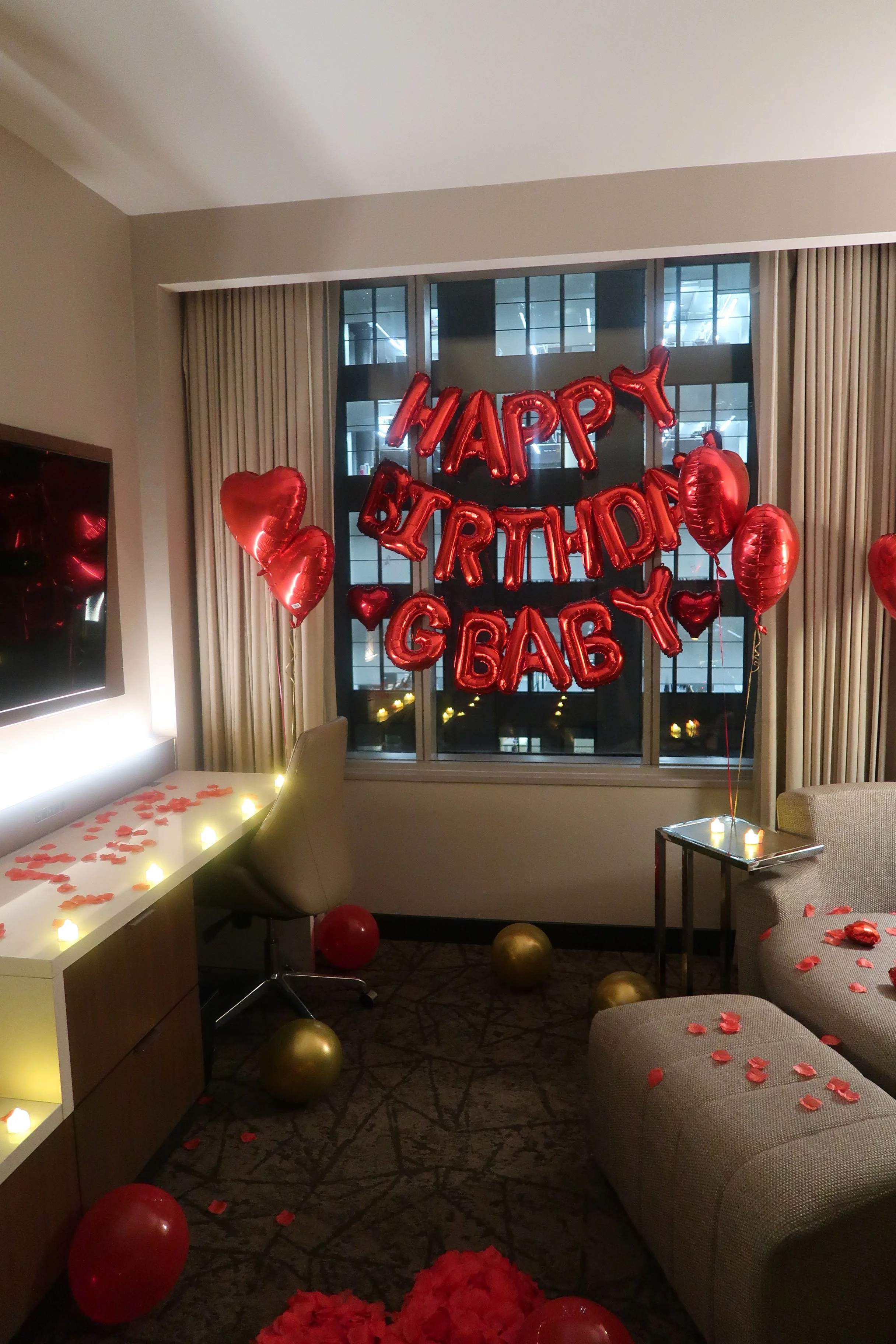 Hotel room decorated with red and gold balloons, 'Happy Birthday GBaby' banner, and rose petals