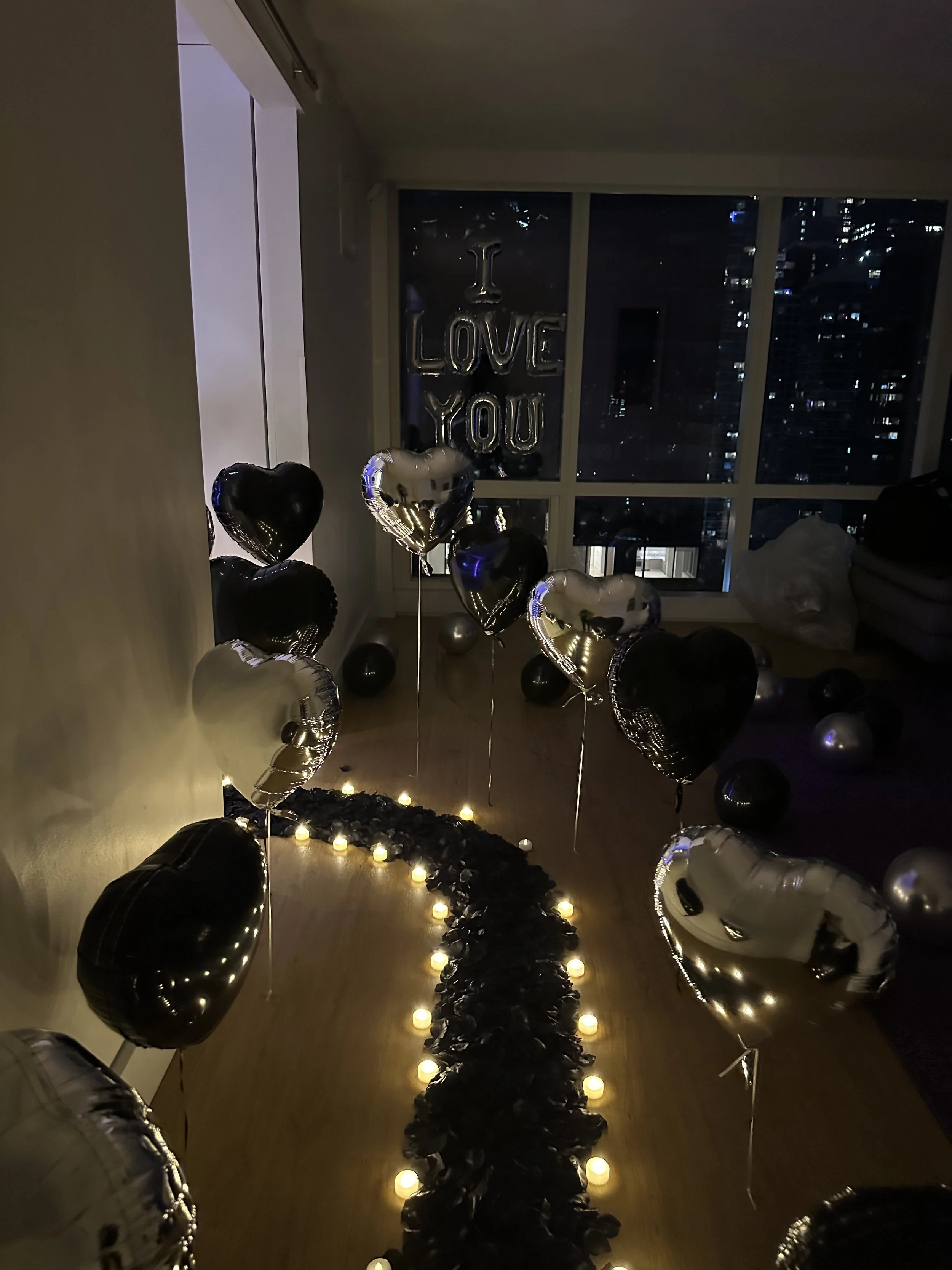 Hotel room decorated with black and silver balloons, 'I Love You' banner, LED candles, and black rose petals