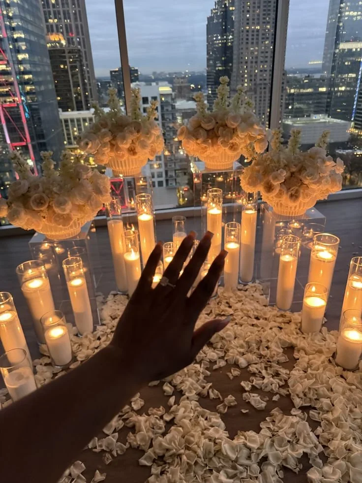 Engagement ring on hand with romantic proposal backdrop featuring elevated floral displays on pillars, candlelight, and scattered rose petals