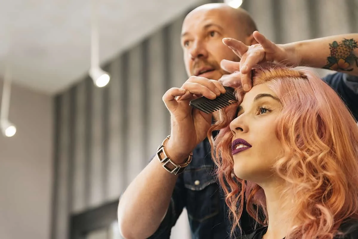 A hairdresser styles a woman's pink curly hair in a salon.