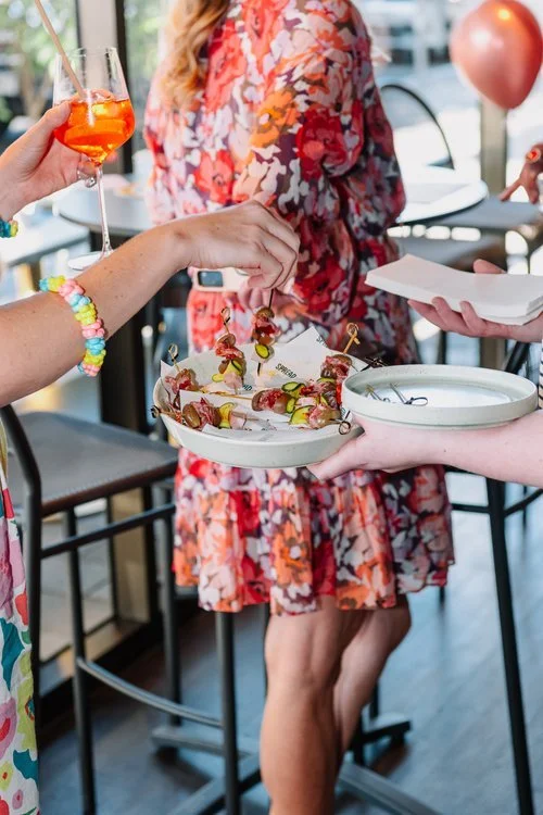 People at a party serving and enjoying drinks and appetizers, including a woman with a colorful wrist bracelet holding a glass of orange cocktail and others serving a plate of food.