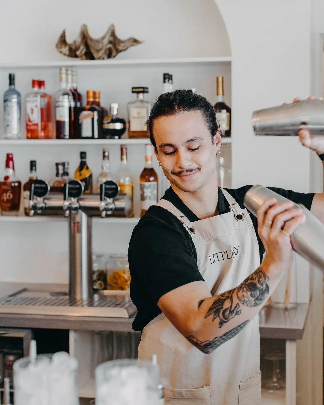 A smiling bartender stirring a cocktail shaker in a bar with bottles of liquor on shelves behind him.