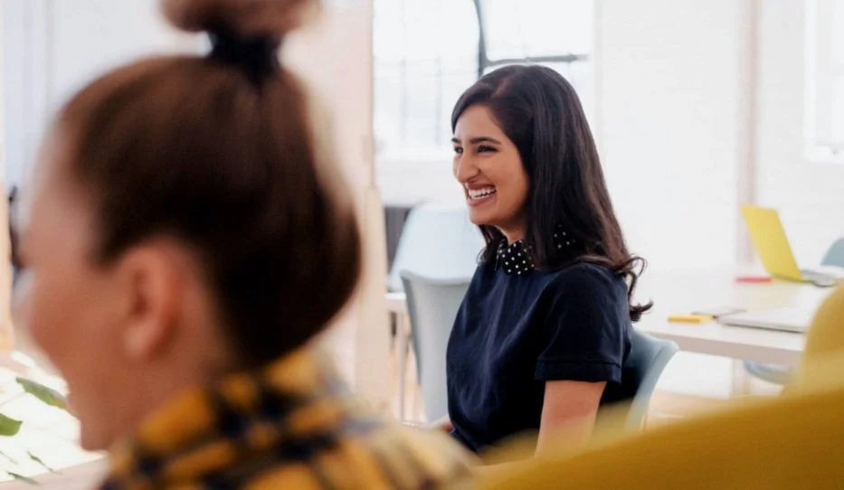 A woman with dark hair, wearing a dark shirt with a collar, is smiling and engaging in conversation with a group of people in a bright, modern office setting.