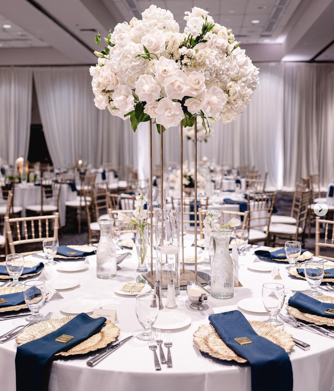 Elegant round table setup for a formal event with a tall floral centerpiece of white roses and hydrangeas, surrounded by gold chiavari chairs. The table is set with blue napkins, gold chargers, glassware, and silverware. The background features drape