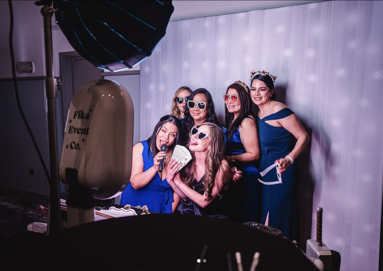 Group of women posing in a photo booth with sunglasses and props like a microphone and fake money.