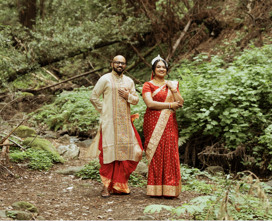A couple dressed in traditional Indian attire standing on a forest trail surrounded by greenery, with the man holding his hand on his chest and the woman holding her arm across her midsection.