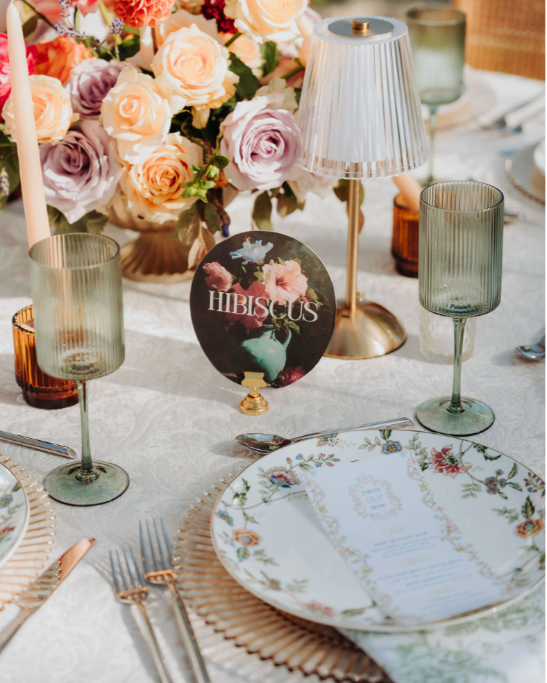 A beautifully set dining table with floral centerpiece, green-tinted glassware, a decorative plate with a floral design, and a small round sign labeled 'HIBISCUS' in front of a large bouquet of roses in pastel colors.