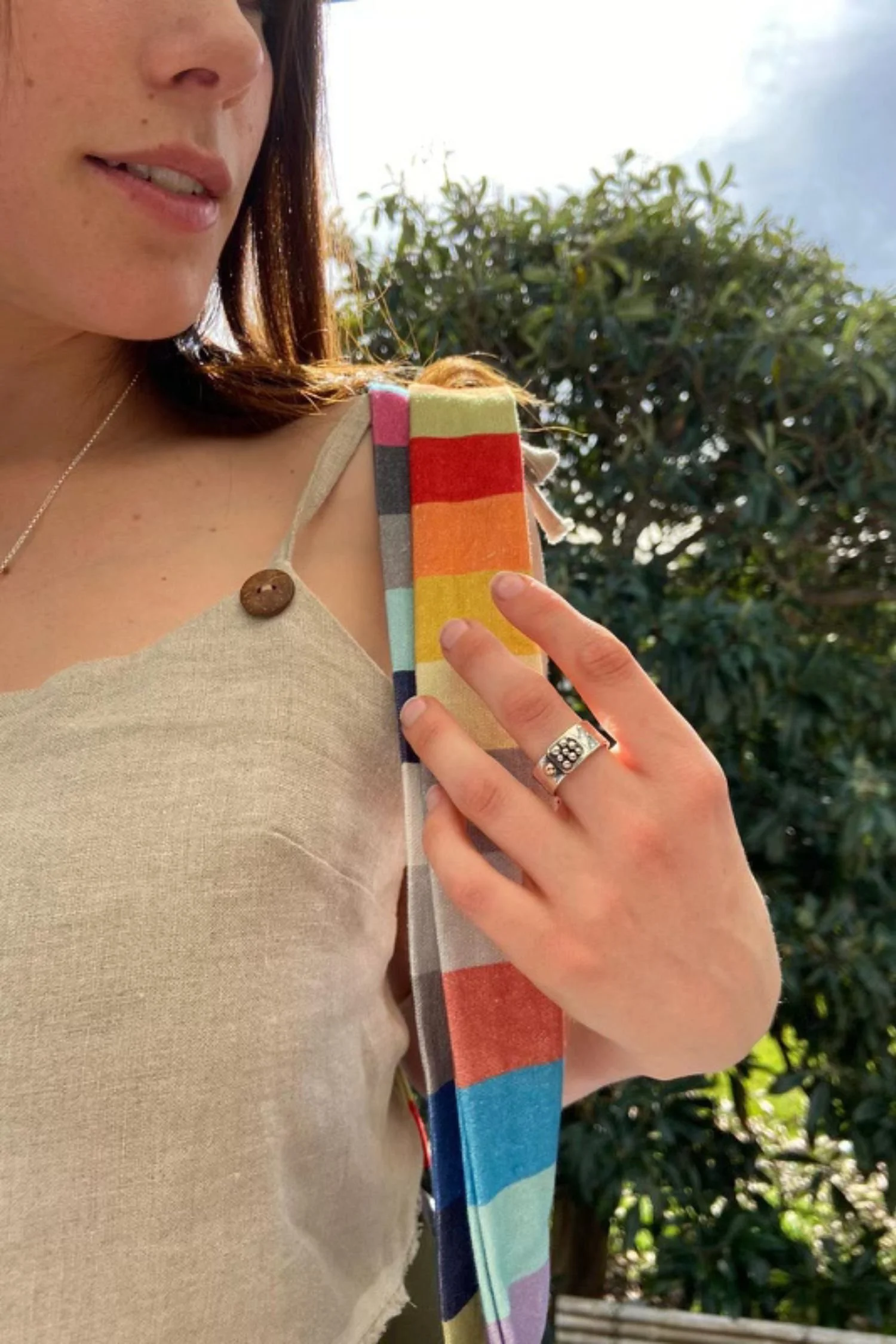 Close-up of a woman wearing a beige top with a wooden button, a silver ring, and a colorful strap with rainbow stripes, standing outdoors near green foliage.