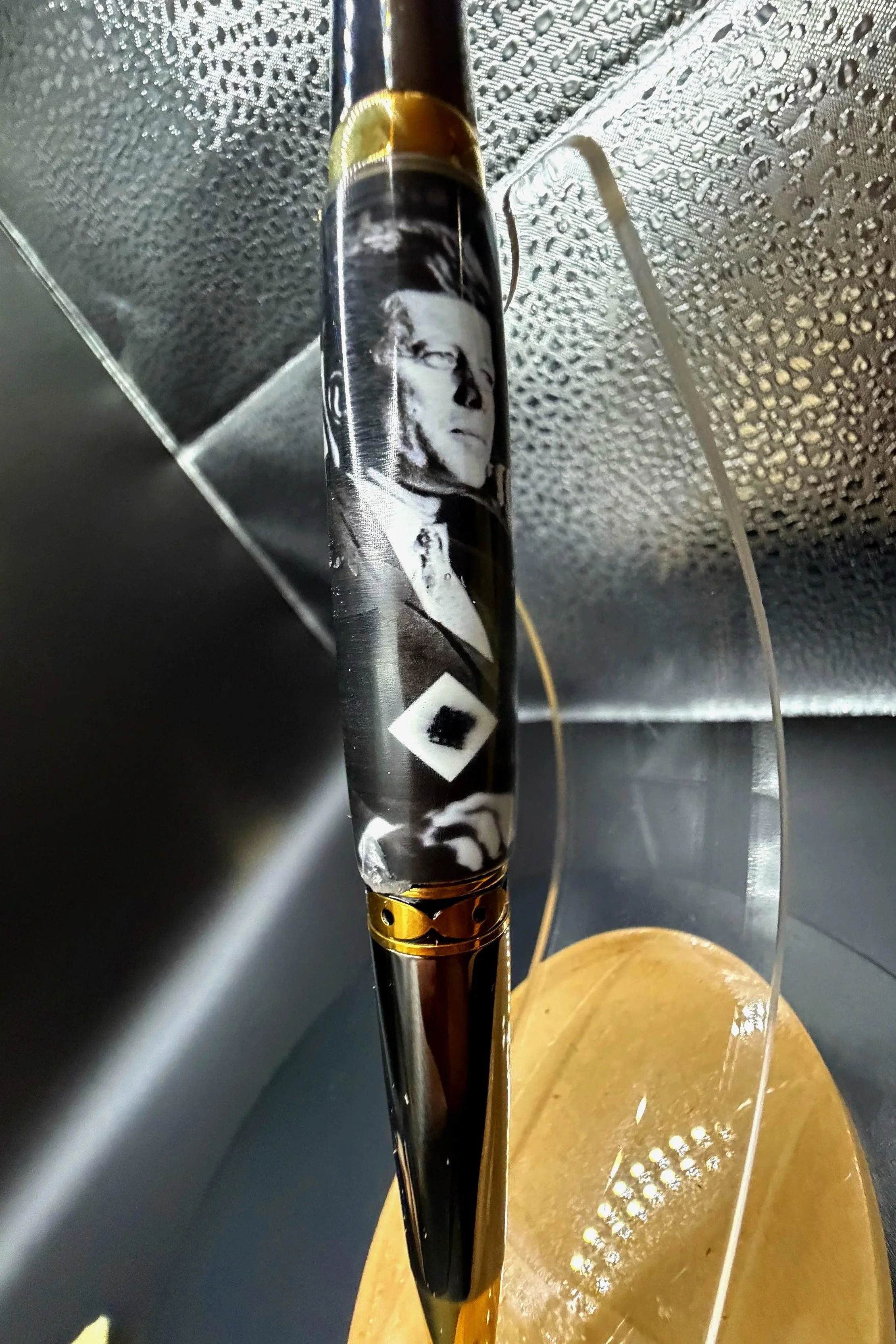 Close-up of a fishing rod with a black and white face image on it, hanging from a ceiling with metallic textured surface, next to a pink and orange curtain.