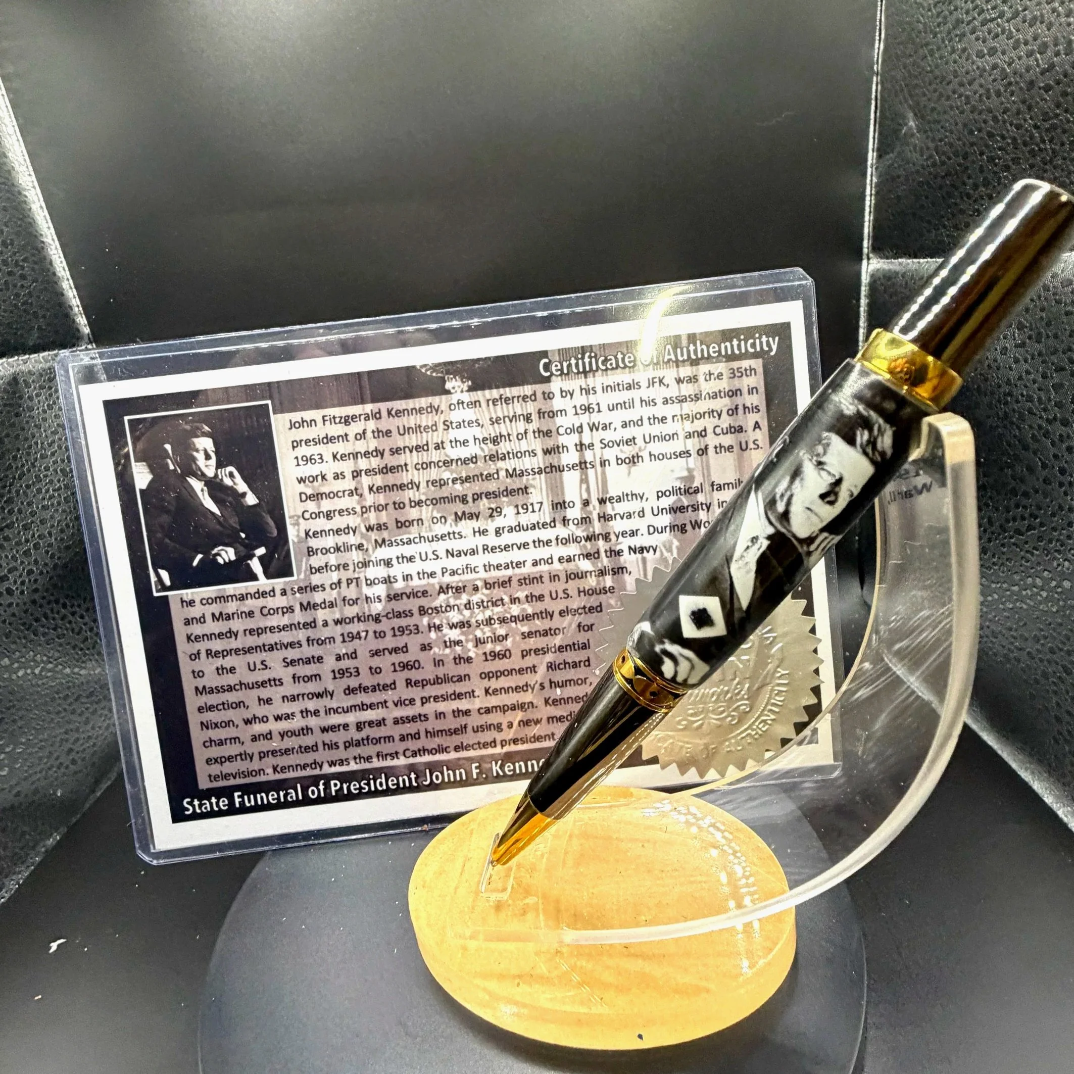 Close-up of a small information card about John F. Kennedy, with black and white photograph and text, next to a decorative glass pen with black and white marbled design, placed on a yellow circular stand.