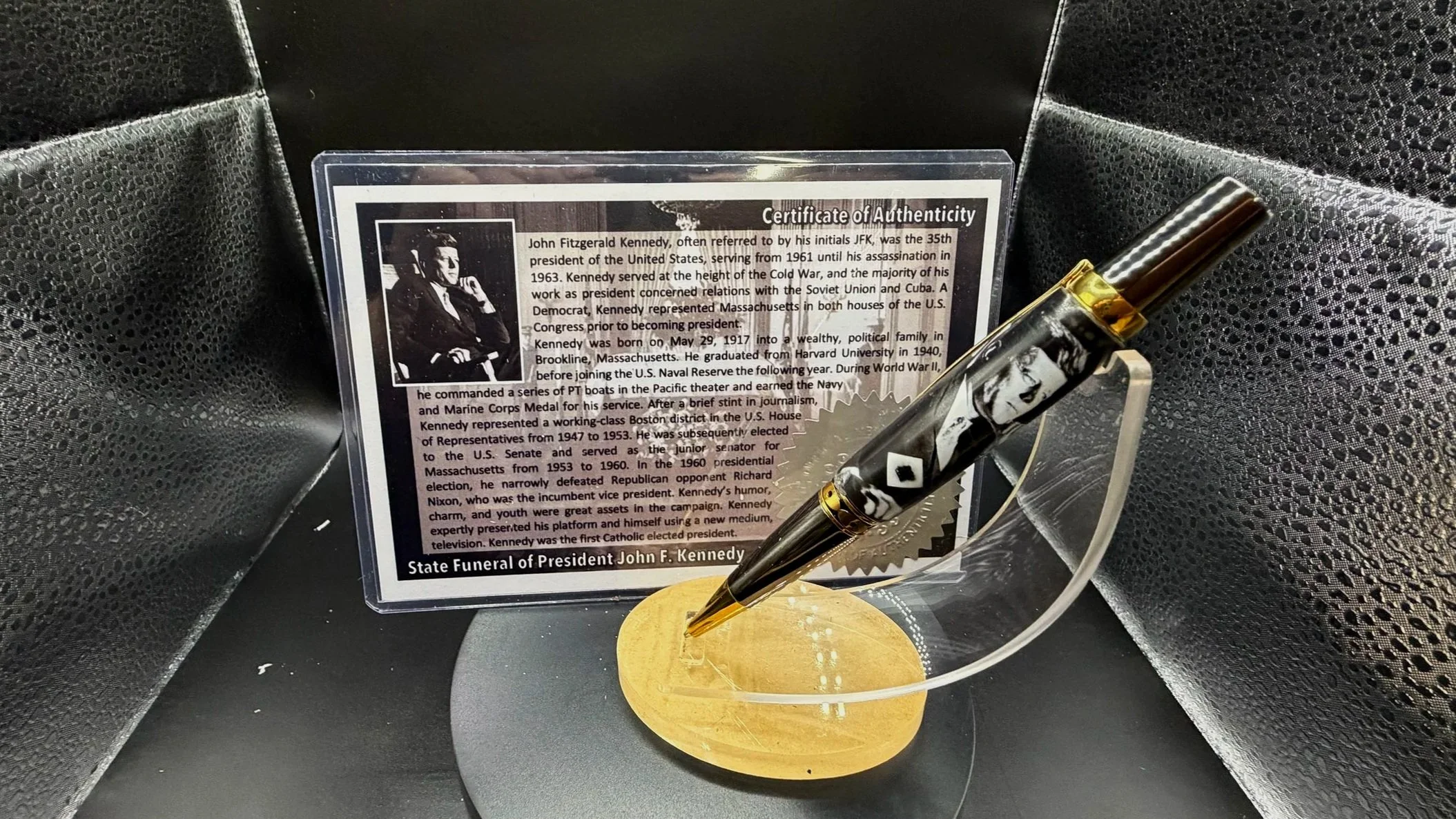 A display with a certificate of authenticity for John F. Kennedy, a commemorative pen, and a gold-colored base, all set against a black textured background.