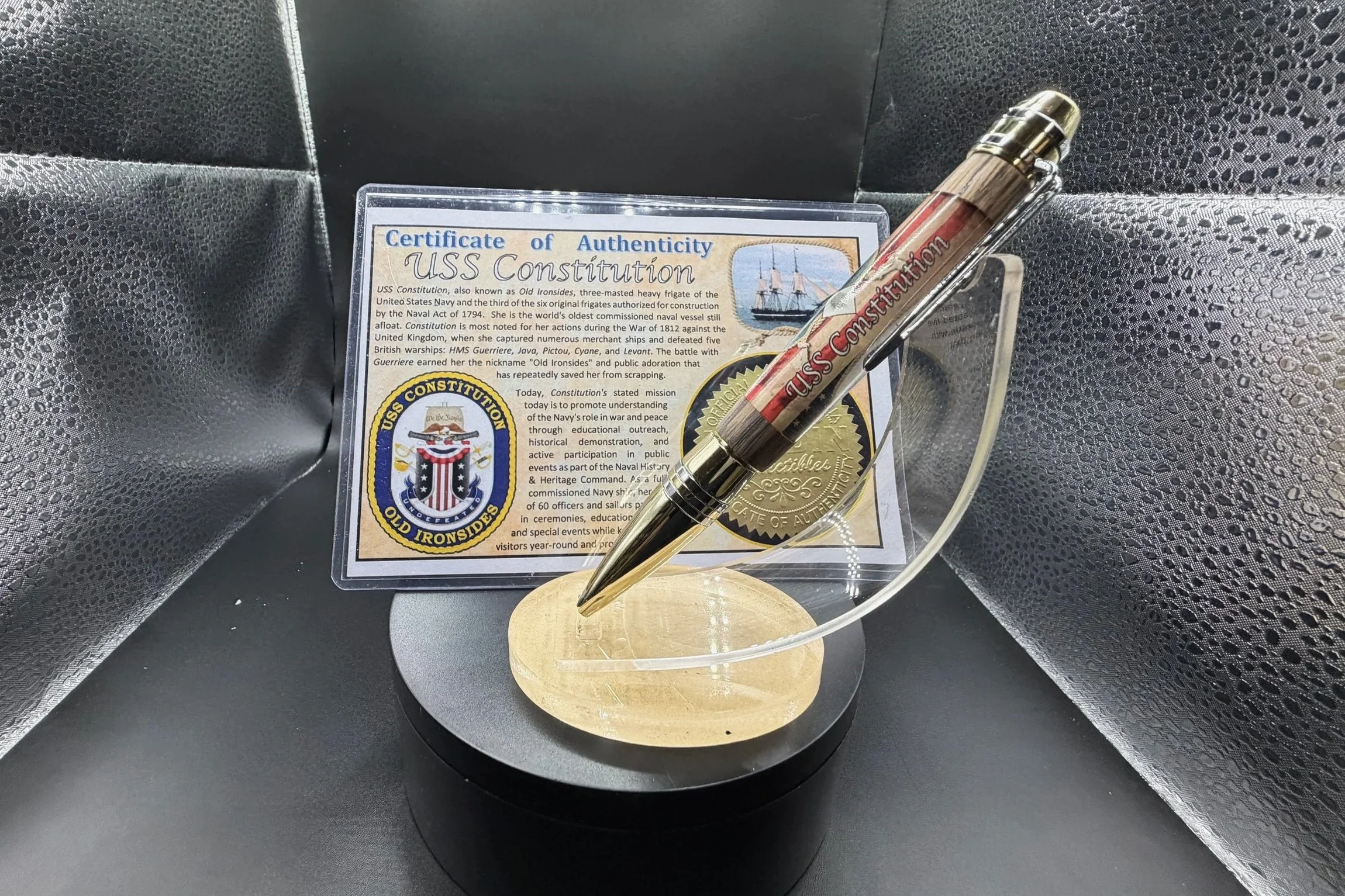 A certificate of authenticity for the USS Constitution, displaying a ship illustration and insignia, along with a pen and a gold medallion, all inside a plastic holder on a black surface with textured black and silver background.