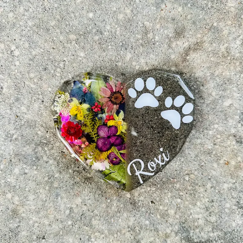 A heart-shaped glass ornament filled with colorful pressed flowers, with paw print designs and the name 'Roxy' on it, placed on a gravel surface.