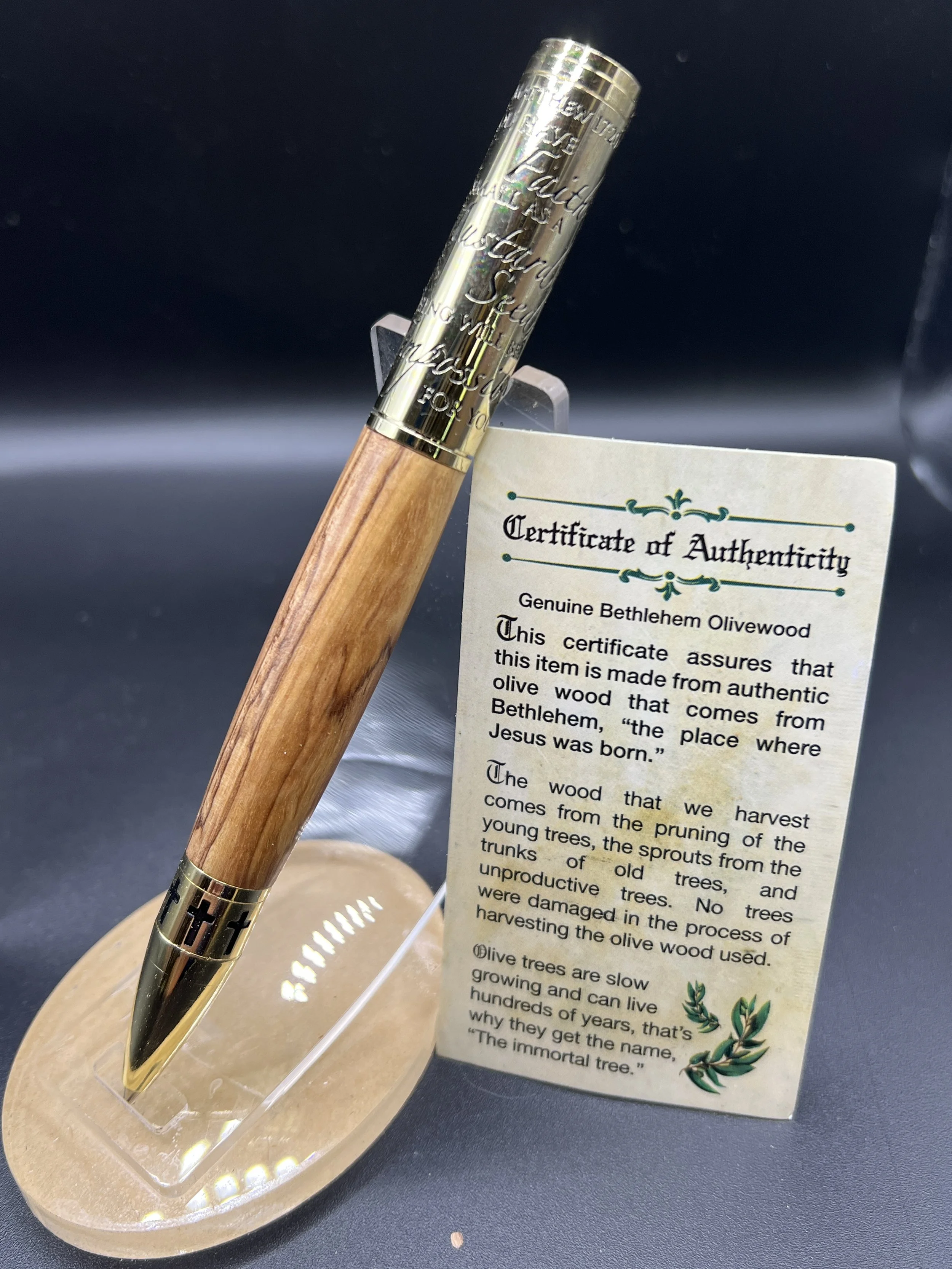 A pen with a gold cap resting on a beige display stand next to a certificate of authenticity for genuine Bethlehem olivewood, with text describing the significance of olive trees and their slow growth.