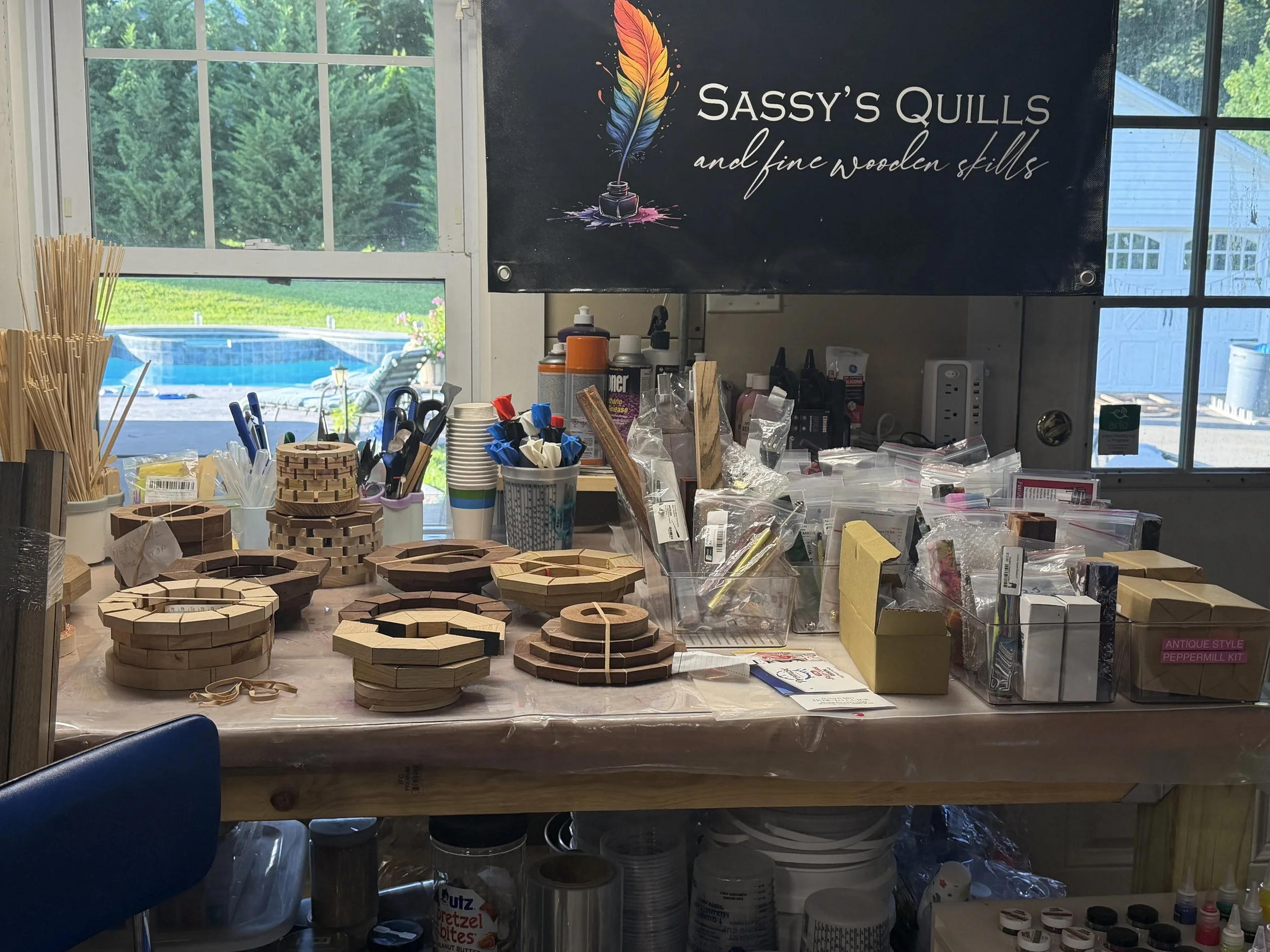 A workshop table with various wooden ring puzzle toys, containers of craft supplies, and tools, with windows showing greenery and a pool outside.