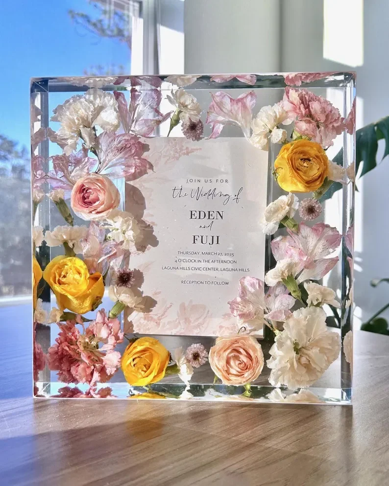 A wedding invitation displayed inside a glass block with floating pink, white, and yellow flowers around the edges, set on a wooden table with a bright window in the background.