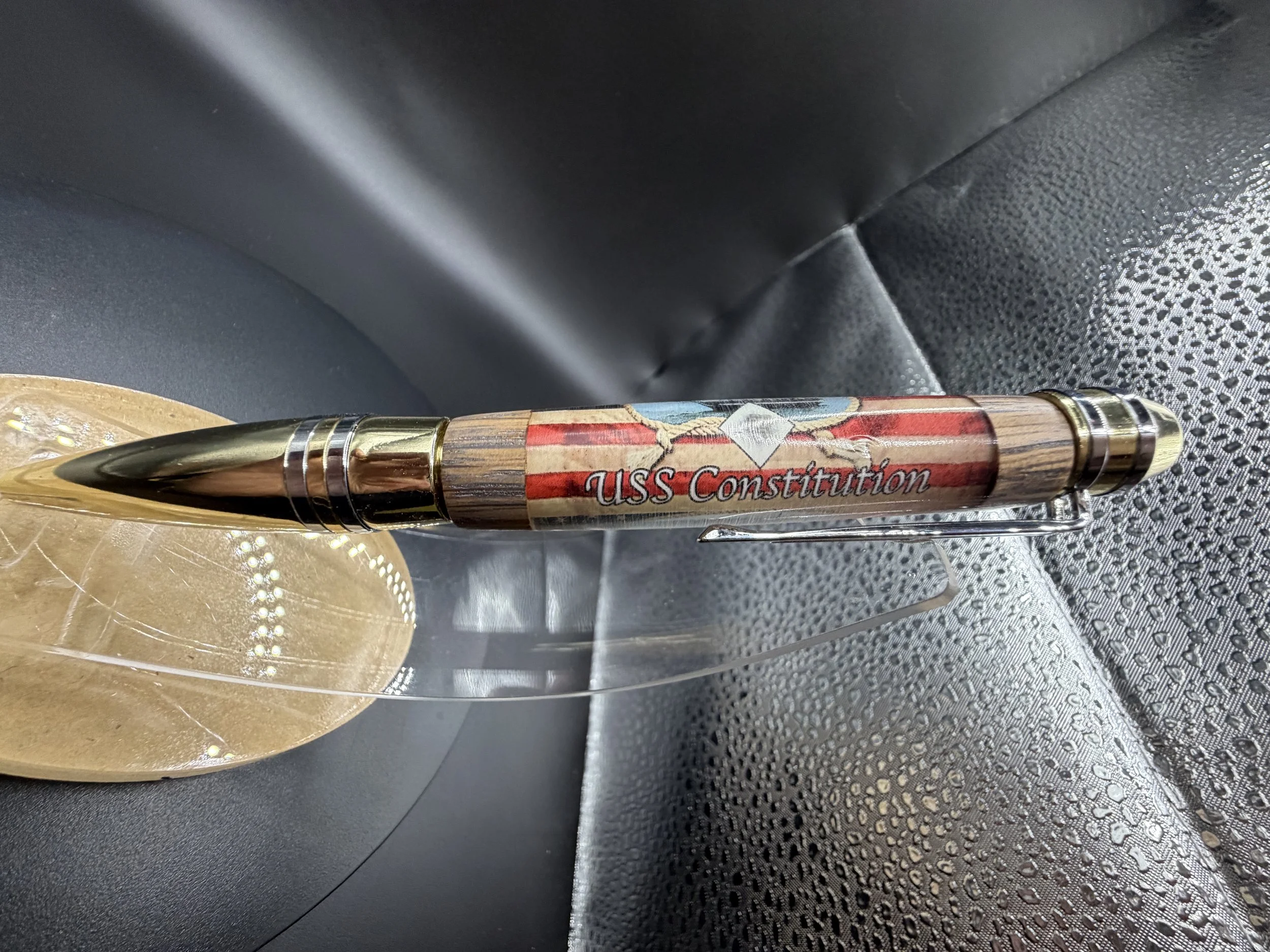 Close-up of a wooden pen with a golden tip and accents, engraved with the words "USS Constitution," resting on a reflective surface with a textured, dark background.