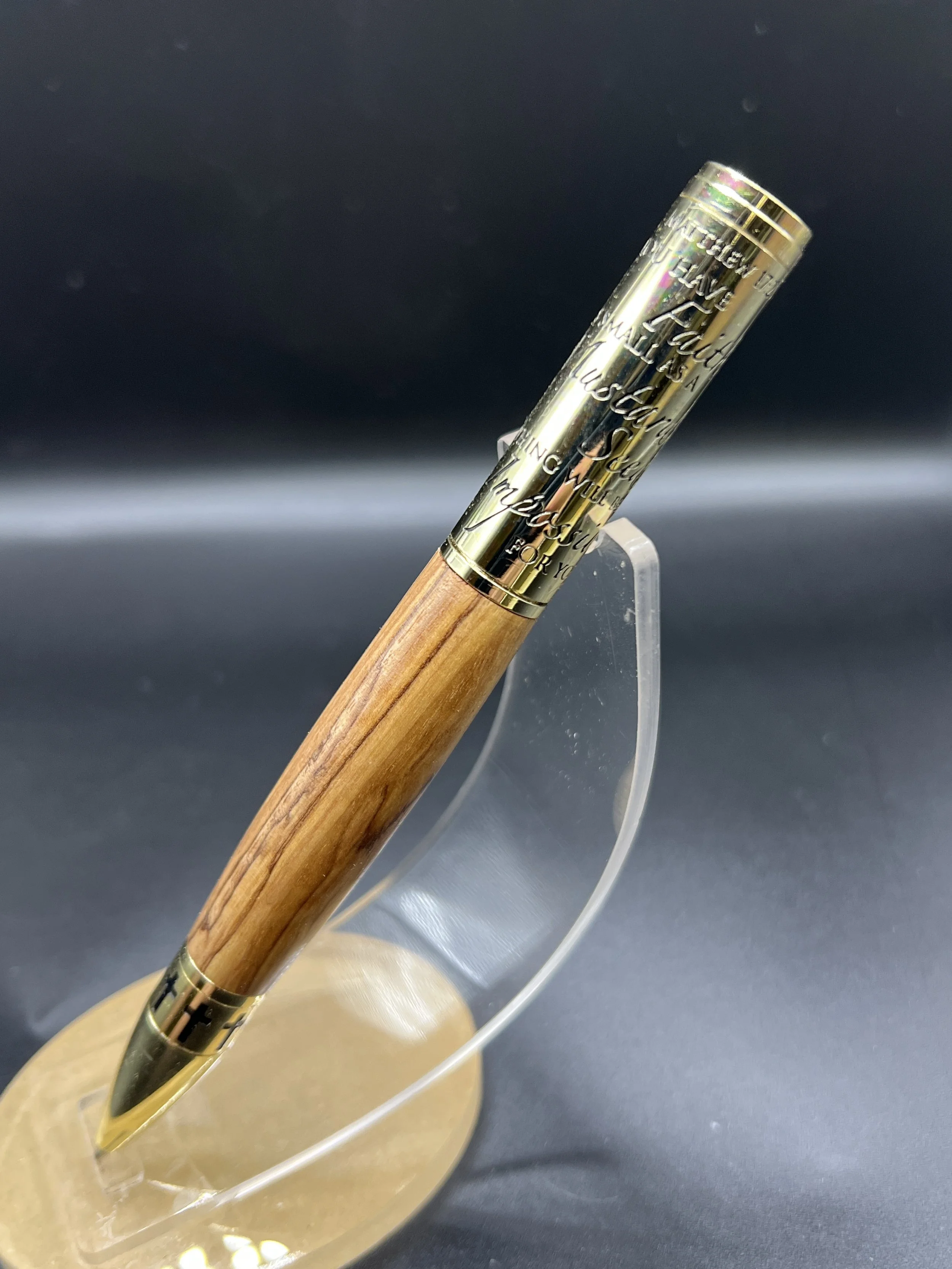 A gold-colored pen with engraved text on its cap, mounted on a transparent stand against a dark background.