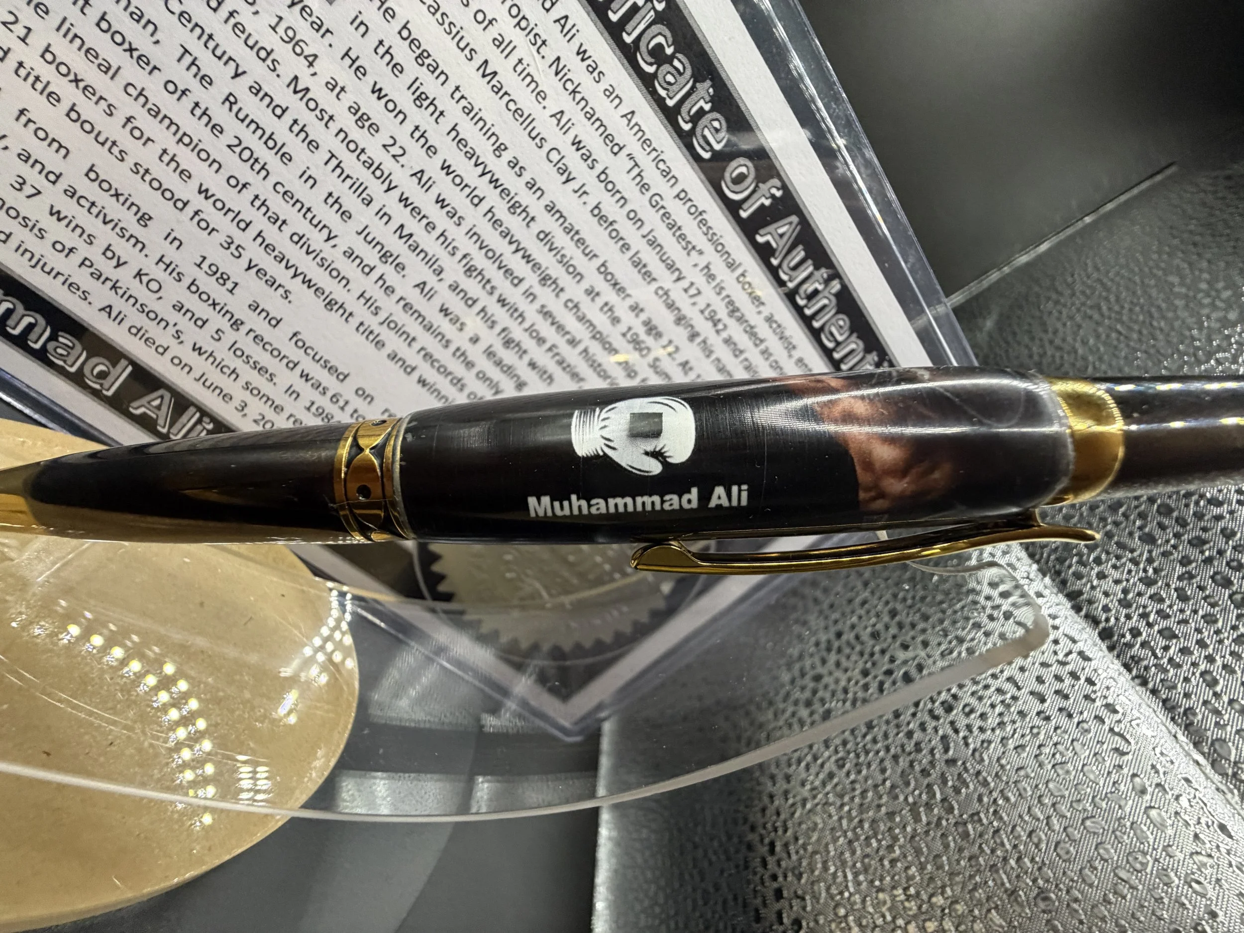 Close-up of a commemorative plaque or trophy with a pen inscribed with 'Muhammad Ali' resting on top, and a decorative gold-colored object beside it. The plaque has detailed text about Muhammad Ali's achievements and history.