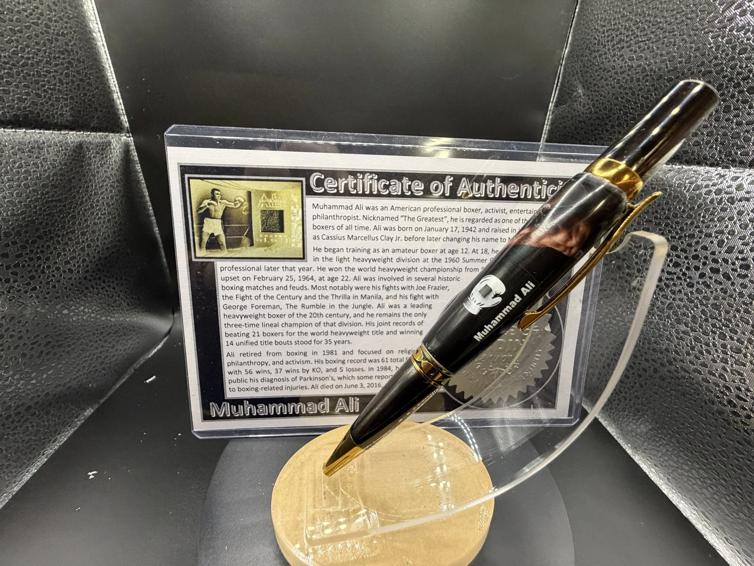 A black pen with gold accents and a clip is resting on a transparent display stand, which holds a printed certificate of authenticity about Muhammad Ali, with a black textured background.
