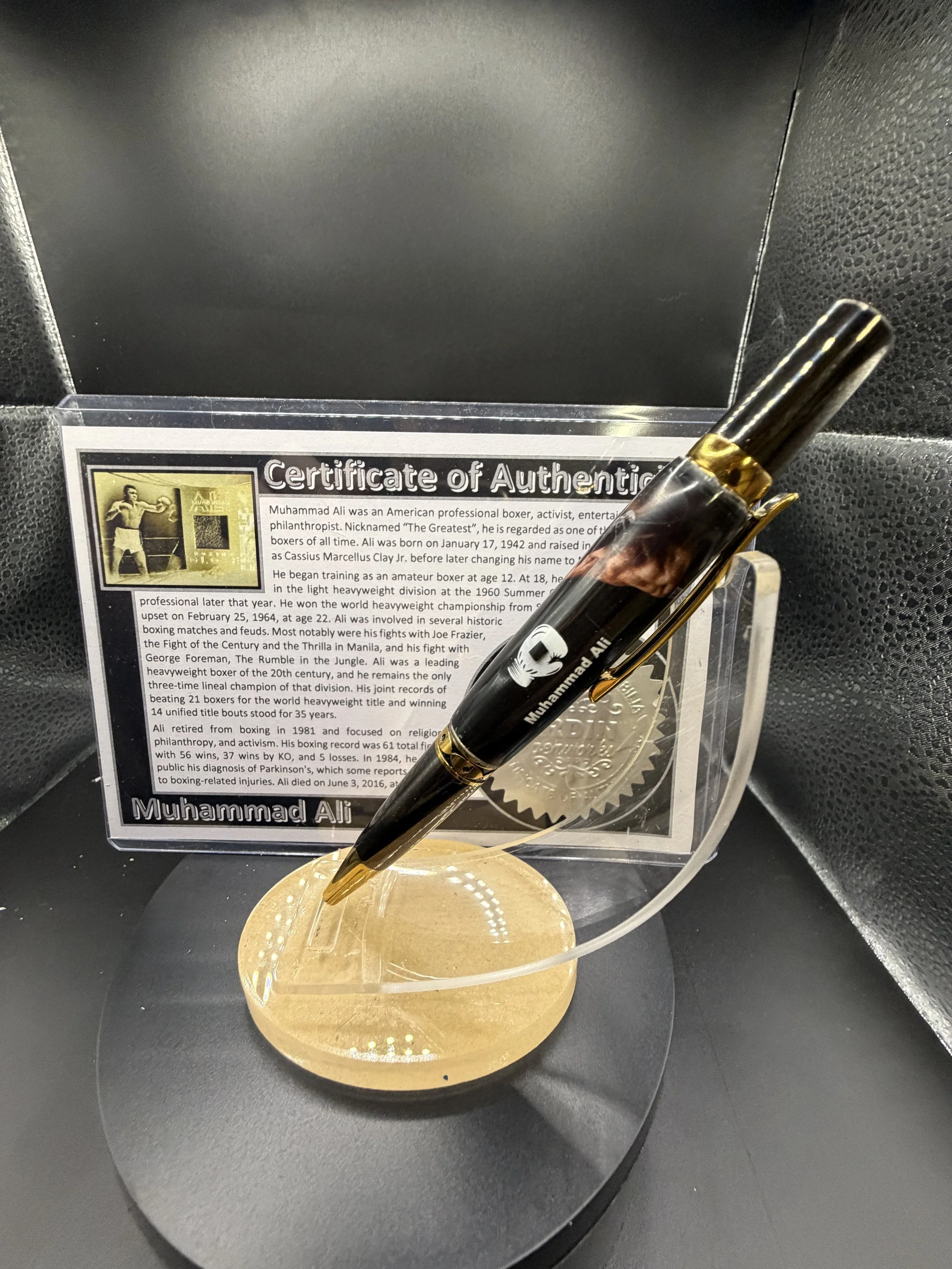 Gold-colored pen with Muhammad Ali's image and signature stands on a circular gold stand, with Muhammad Ali's certificate of authenticity and a silver medal behind it.