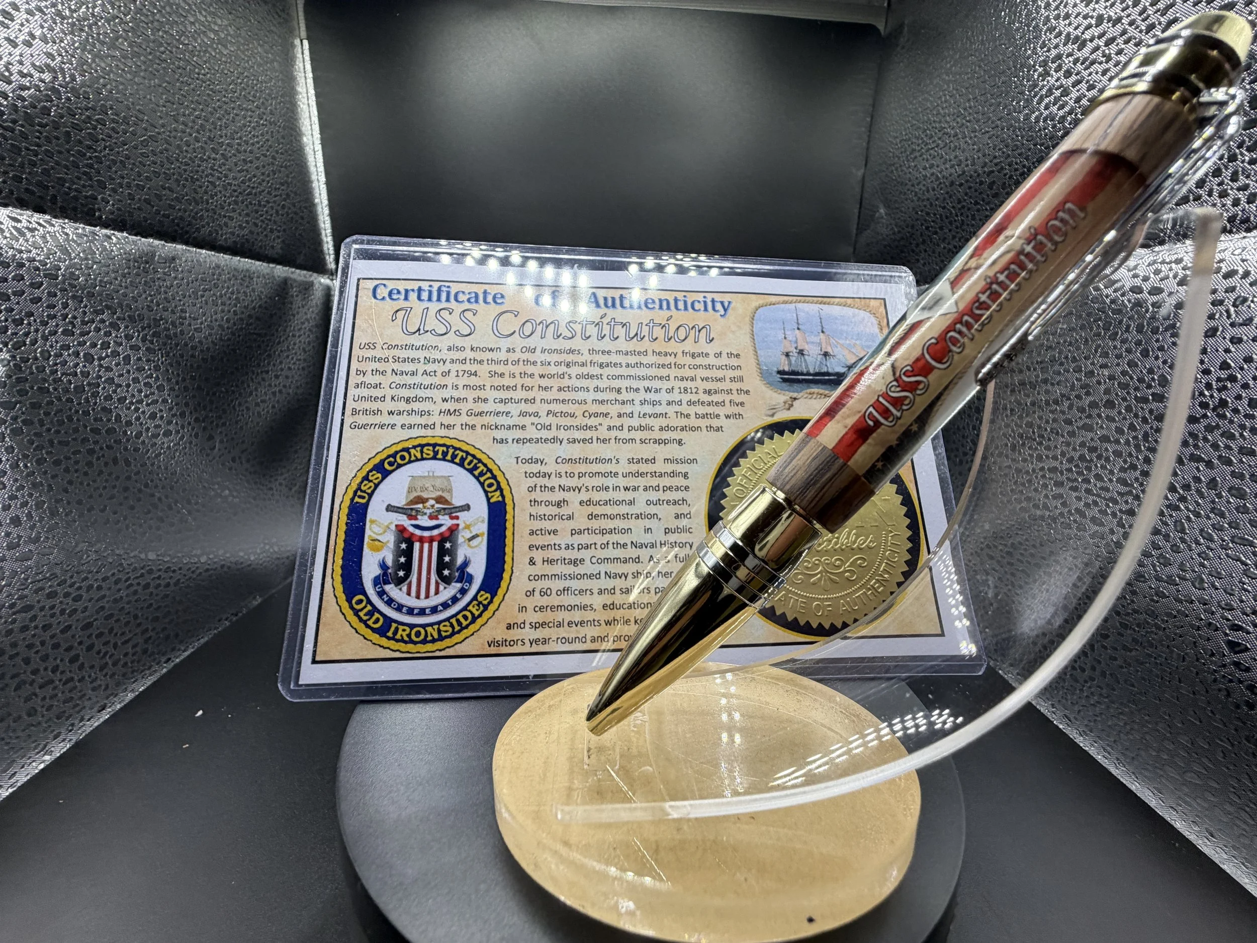 A display of a certificate titled USS Constitution, a gold-colored medal, and a pen resting on a stand with a black and silver textured background.
