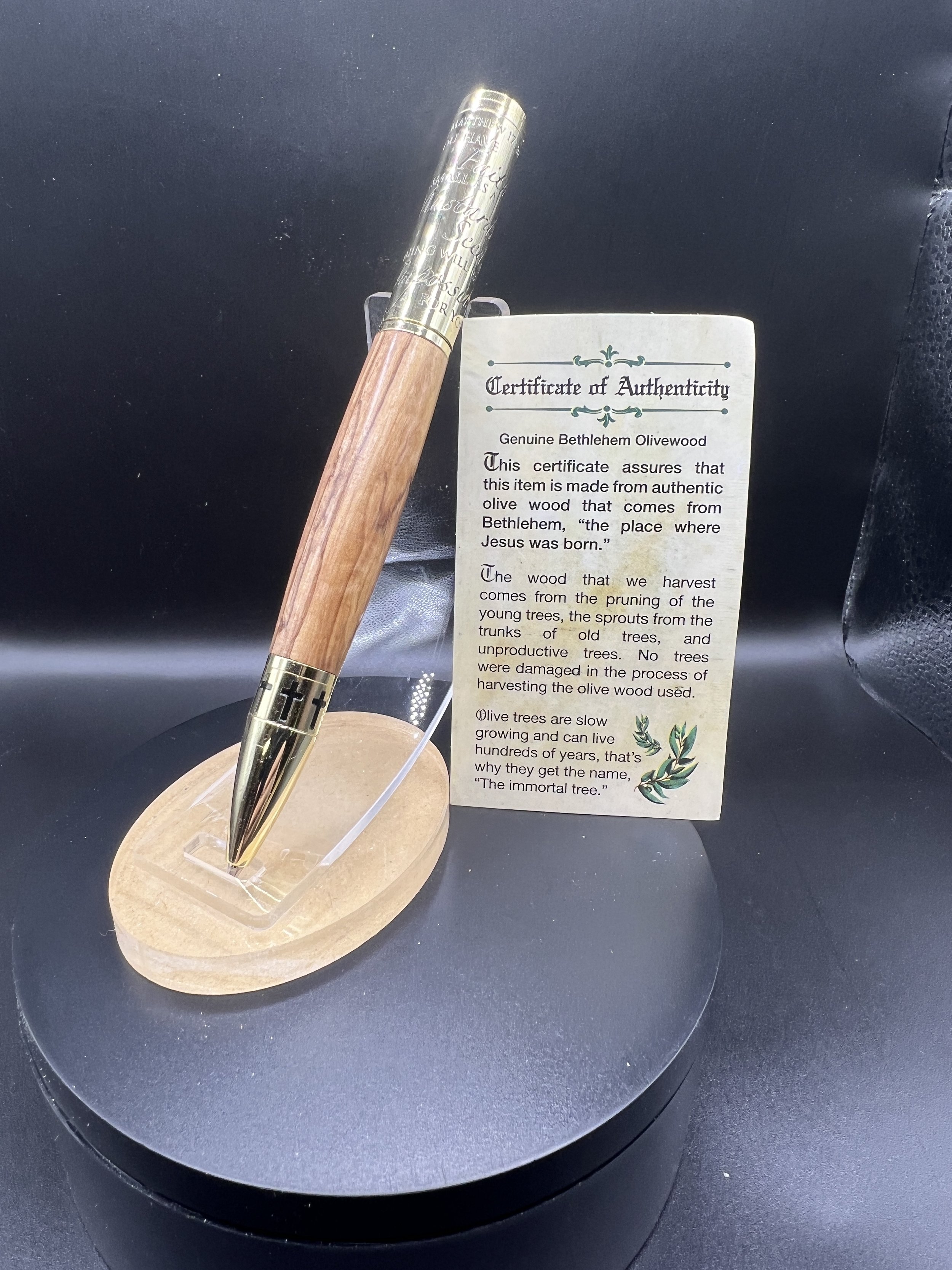 A wooden-handled pen with gold accents and engraved text, displayed on a wooden stand, with a certificate of authenticity for a piece of Bethlehem Olivewood beside it.