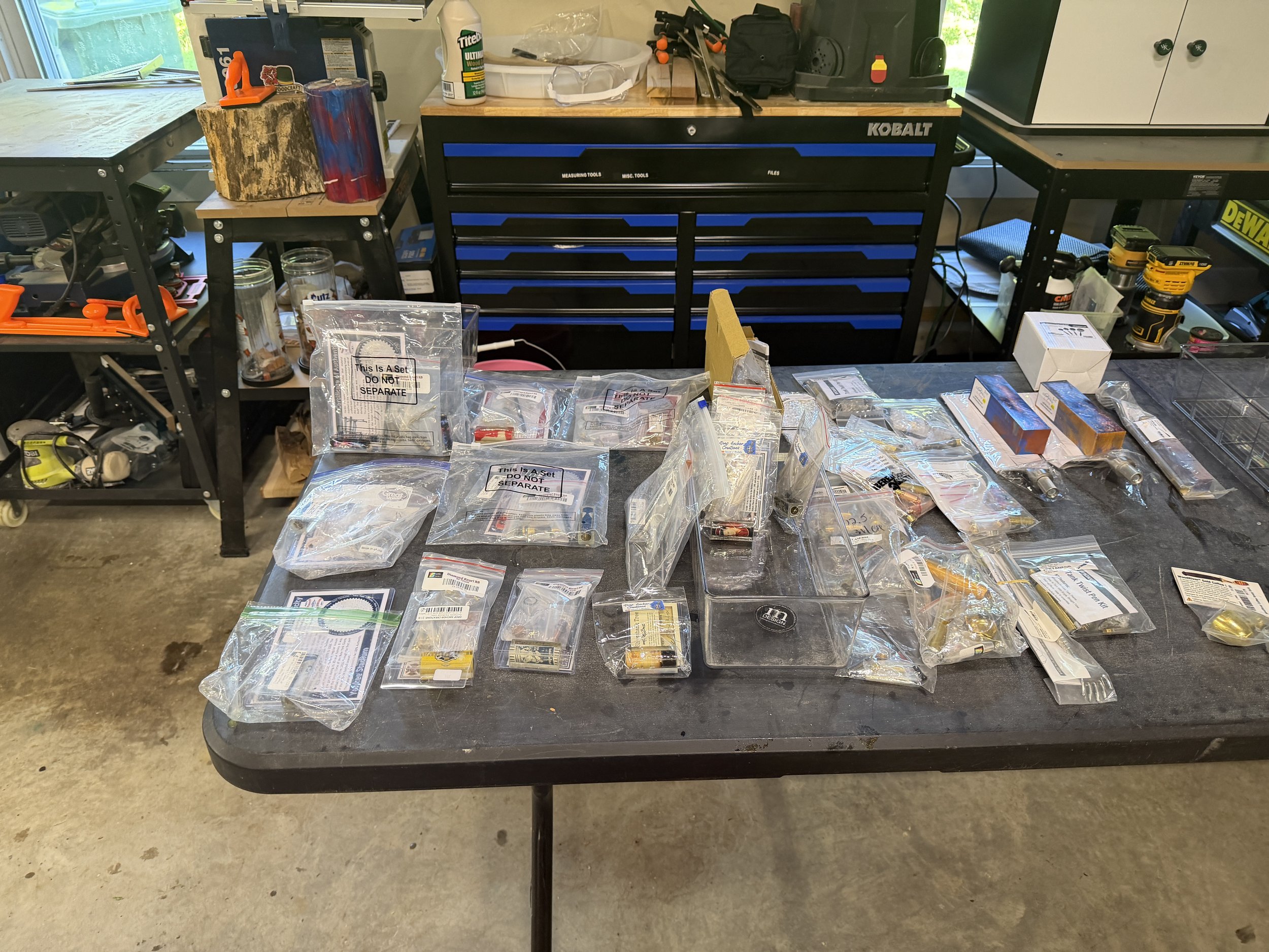 A cluttered black workbench with plastic bags containing small tools and parts, a yellow power drill, a white box, and various other tools. Behind the workbench, there are additional tools and equipment, including a Kobalt tool chest, a window, and s