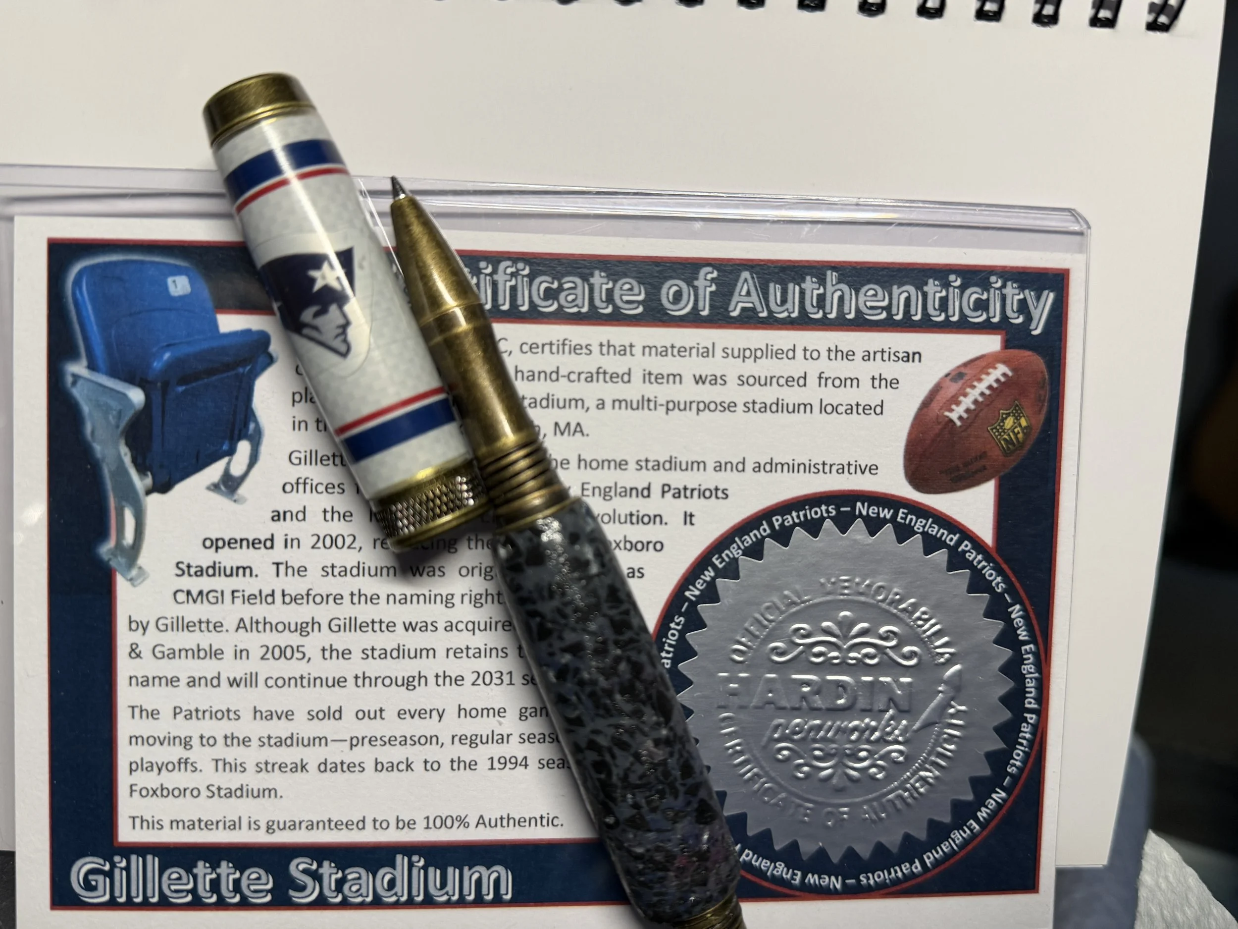 Close-up of a certificate of authenticity for a handcrafted item, with two pens resting on top, partially covering the document. The certificate features a blue, red, and white color scheme and images of sports memorabilia, including a football and a