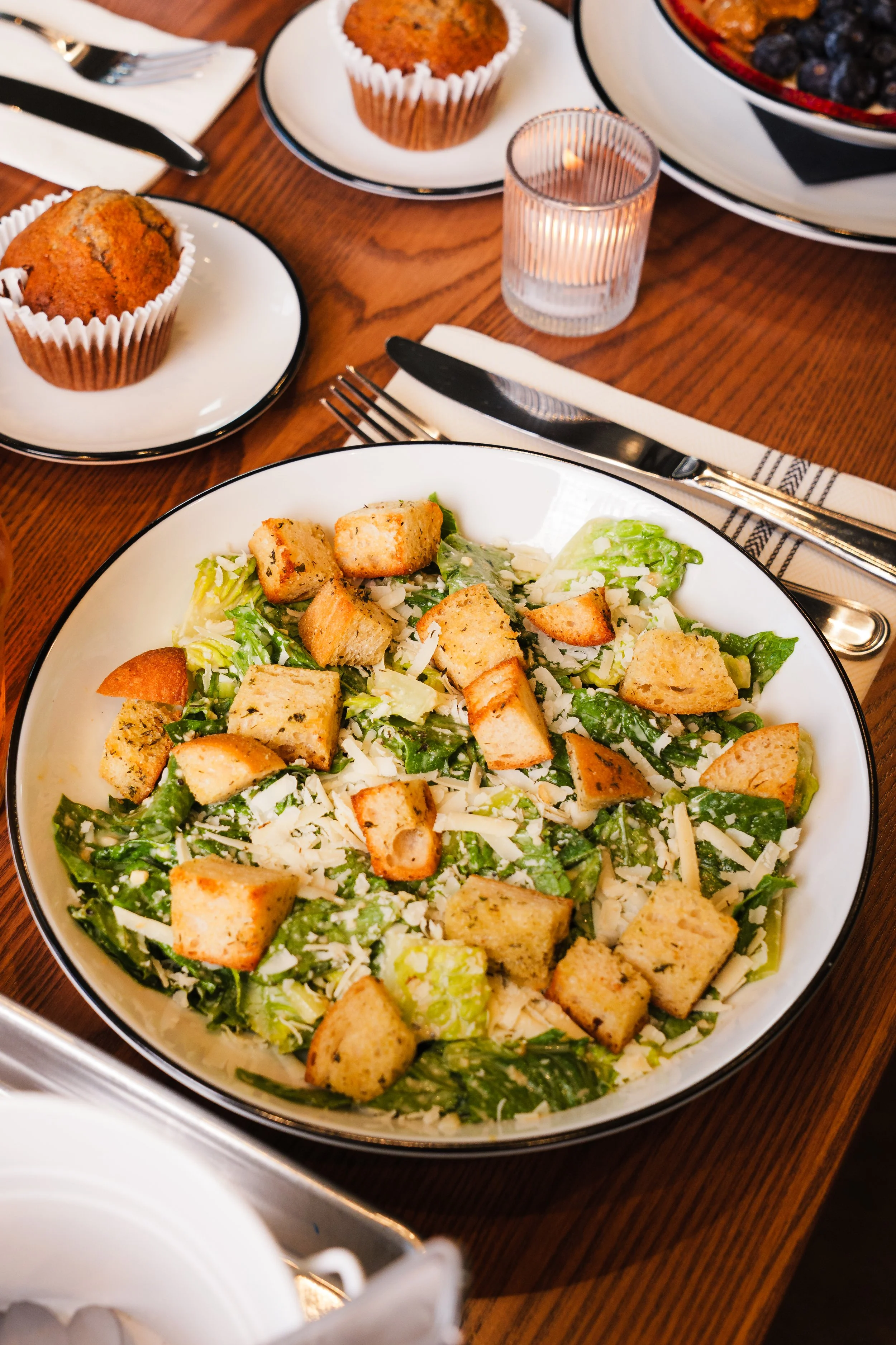 A Caesar salad with croutons and shredded cheese on a white plate, surrounded by muffins, a bowl of berries, utensils, and a lit candle on a wooden table.