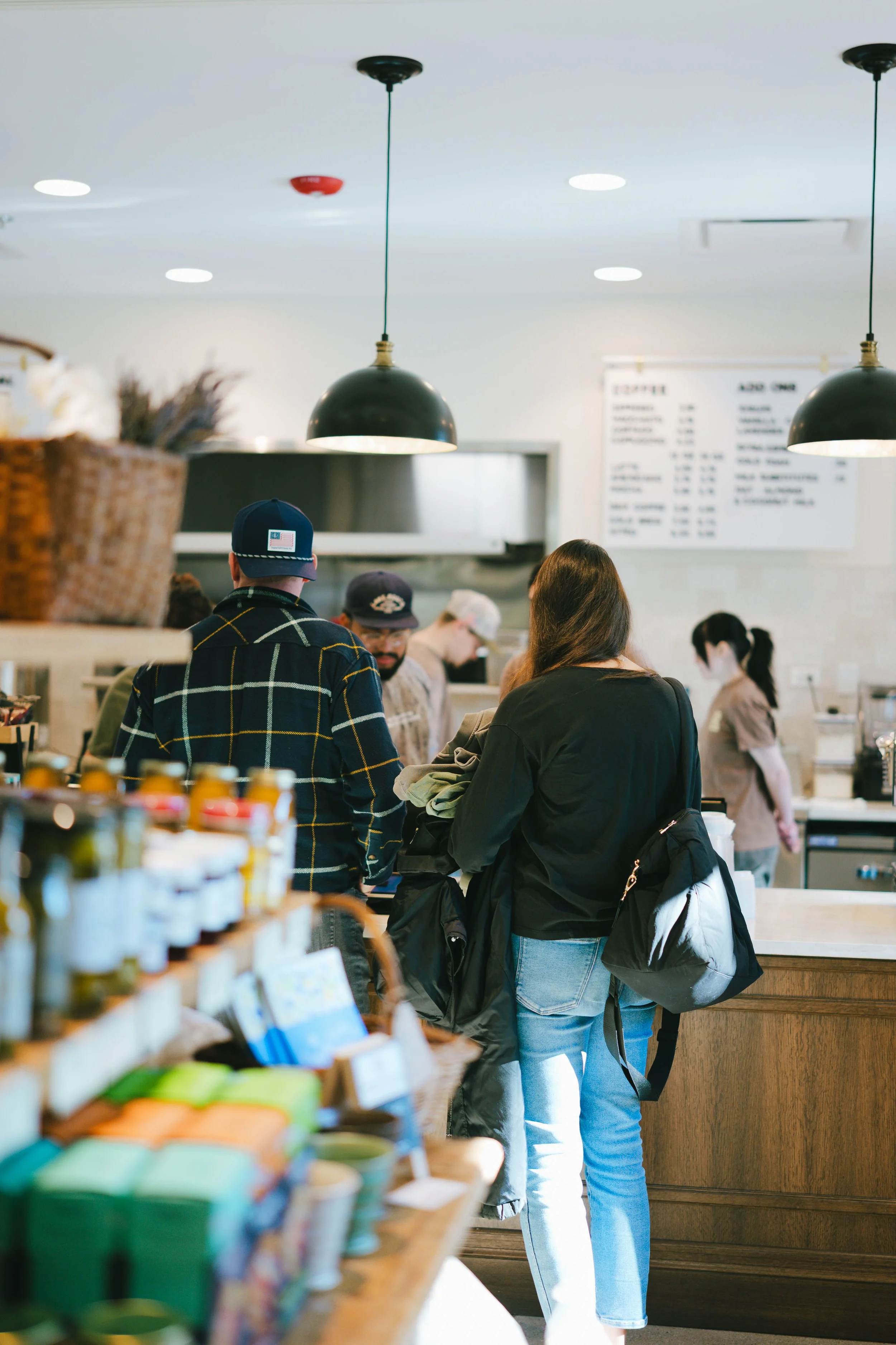 People ordering at a coffee shop counter, with menu board and kitchen in background, and shelves with jars and products in foreground.