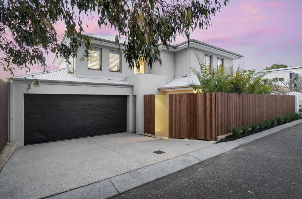 Modern two-story house with a dark garage door, surrounded by a wooden fence and landscaping, under a pink sky at dusk.