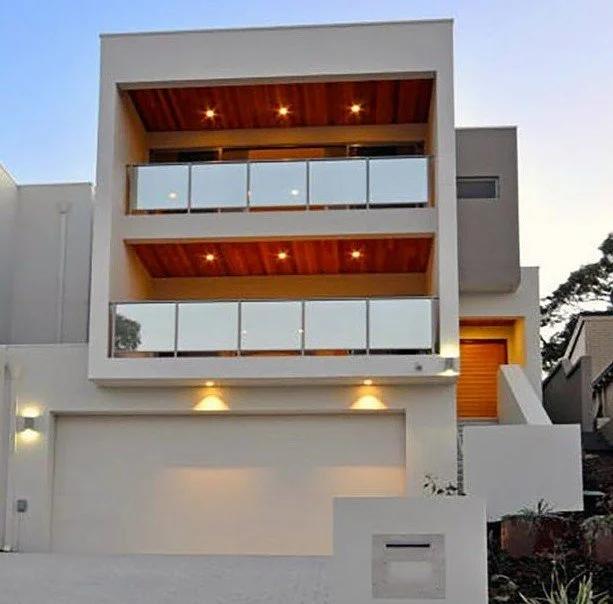 Modern three-story house with large balconies, glass railings, and a garage, illuminated by exterior lights.