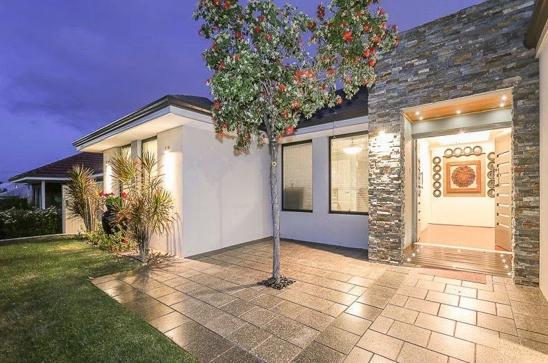 Modern home exterior with stone accents, lit entryway, single tree, landscaped yard, and tiled pathway.