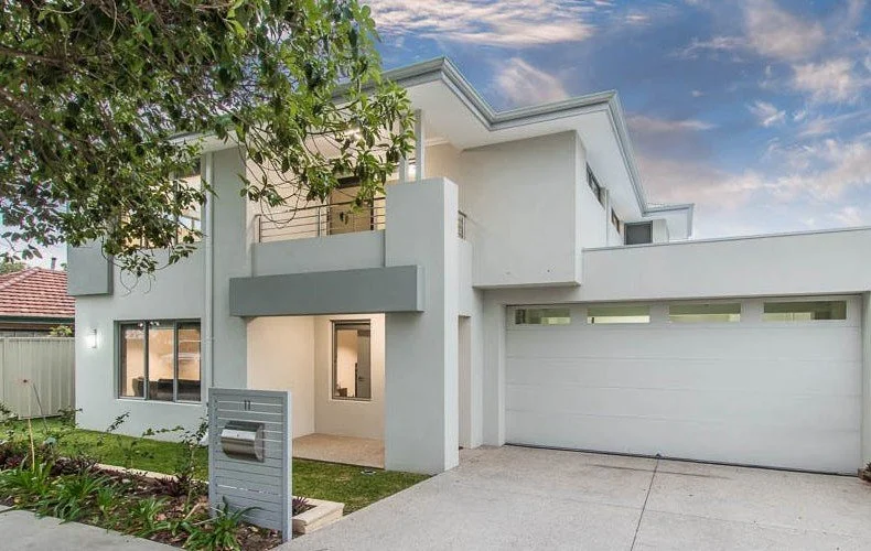 Modern two-story house with flat roof, garage, large windows, and a small front yard under a partially cloudy sky.