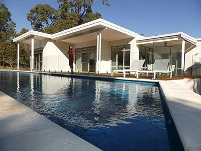 Modern white house with a swimming pool and deck chairs, set against a blue sky and surrounded by trees.
