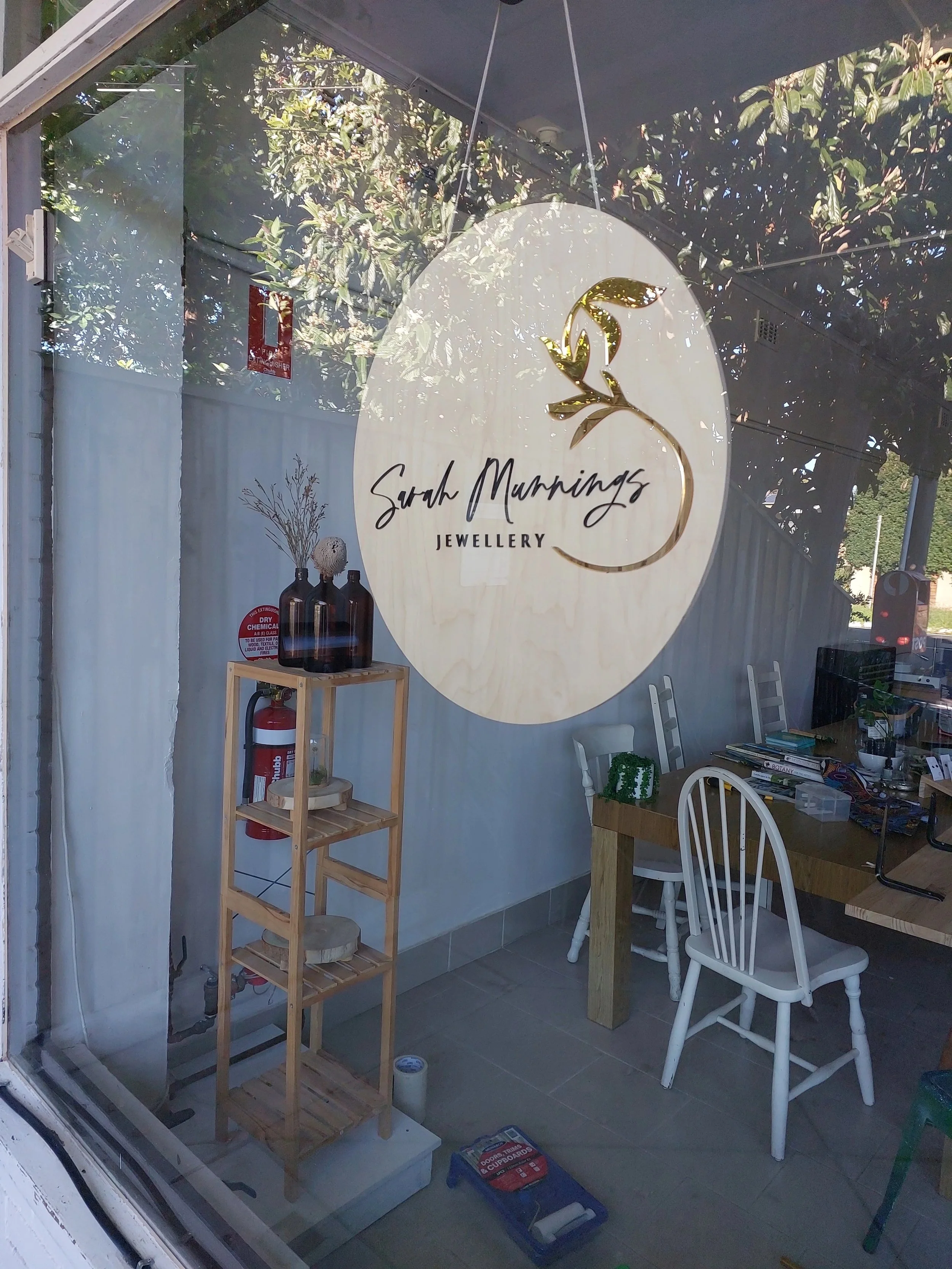 Interior view of Sarah Murnings Jewellery store with a large round logo on the window, showing a gold floral design and the store's name, white chairs, a wooden table with jewelry supplies and books, and a small wooden display shelf with decorative bottles and a fire extinguisher.